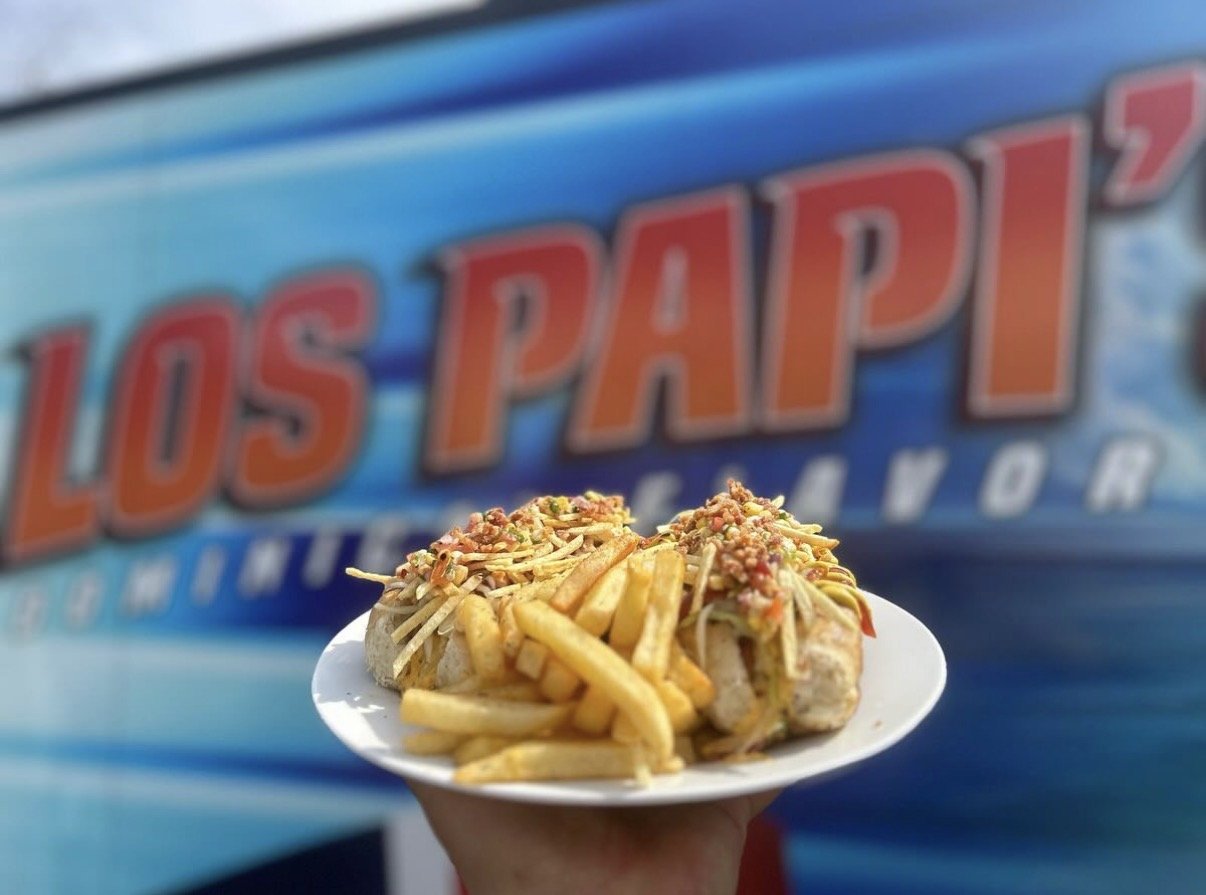 A person holding a plate with a hot dog topped with French fries and crushed chips, in front of a blue sign that says "Los Papis."