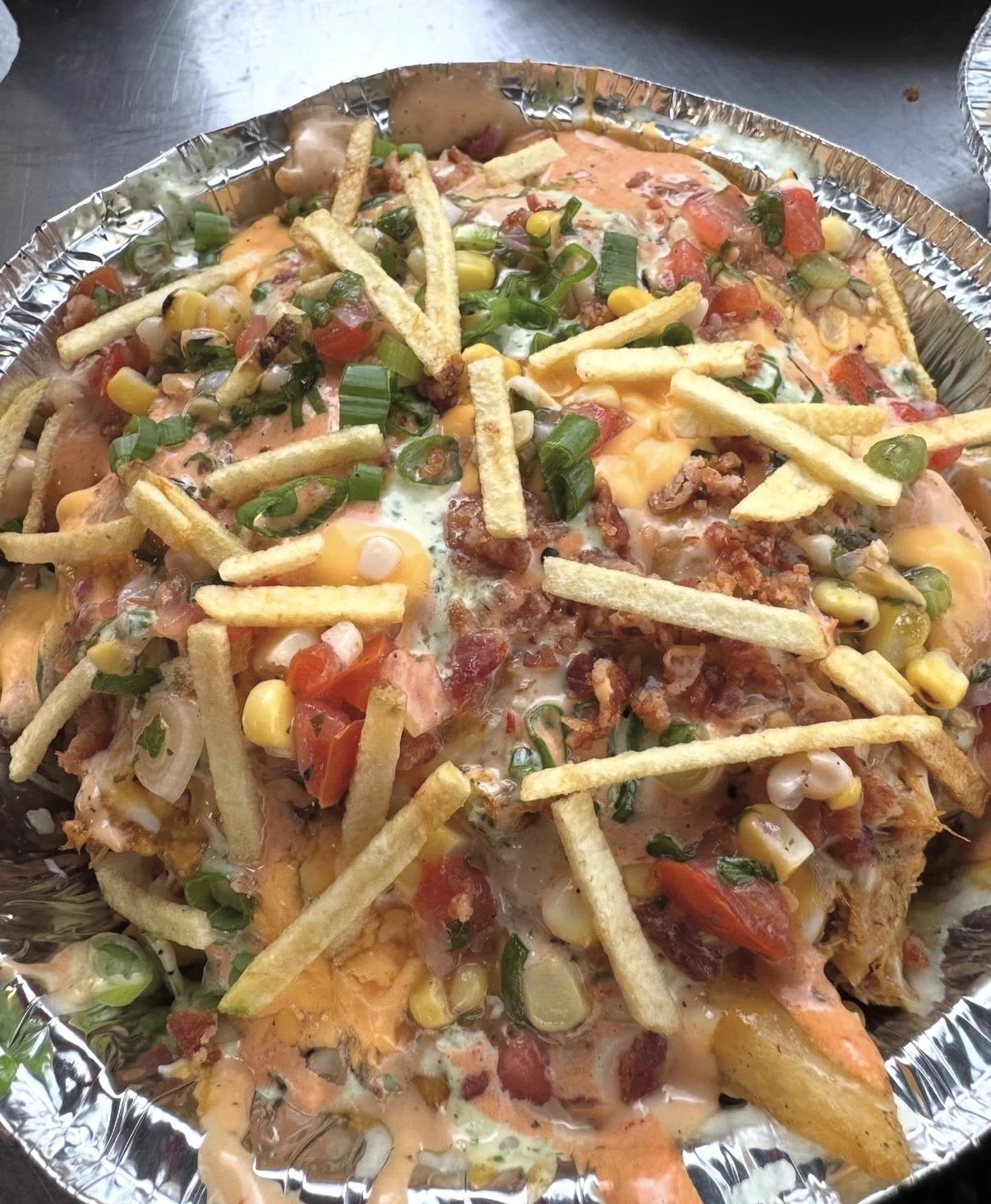 Loaded nachos topped with cheese, chopped tomatoes, green onions, sour cream, and French fries in a foil container.