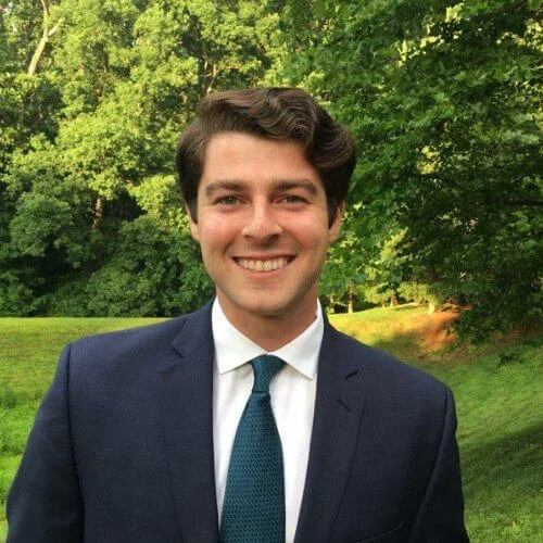 A young man in a navy suit, white shirt, and teal tie standing outdoors in a green park with trees in the background, smiling at the camera.