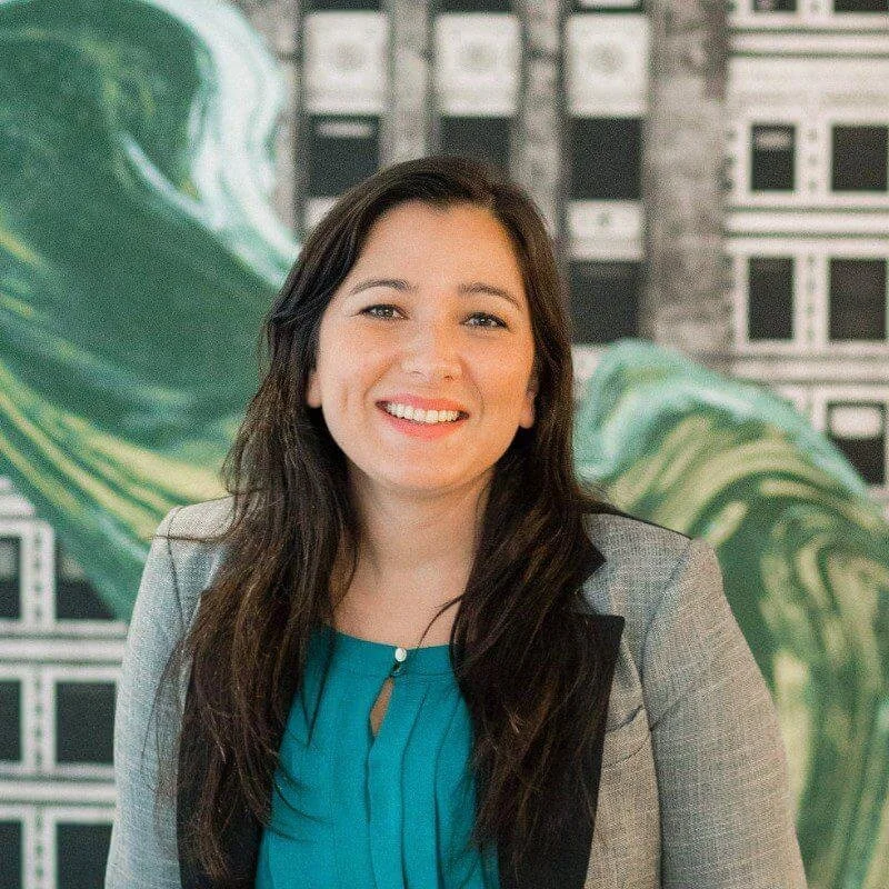 A smiling young woman with long dark hair, wearing a gray blazer over a teal blouse, standing outdoors with a colorful mural in the background.