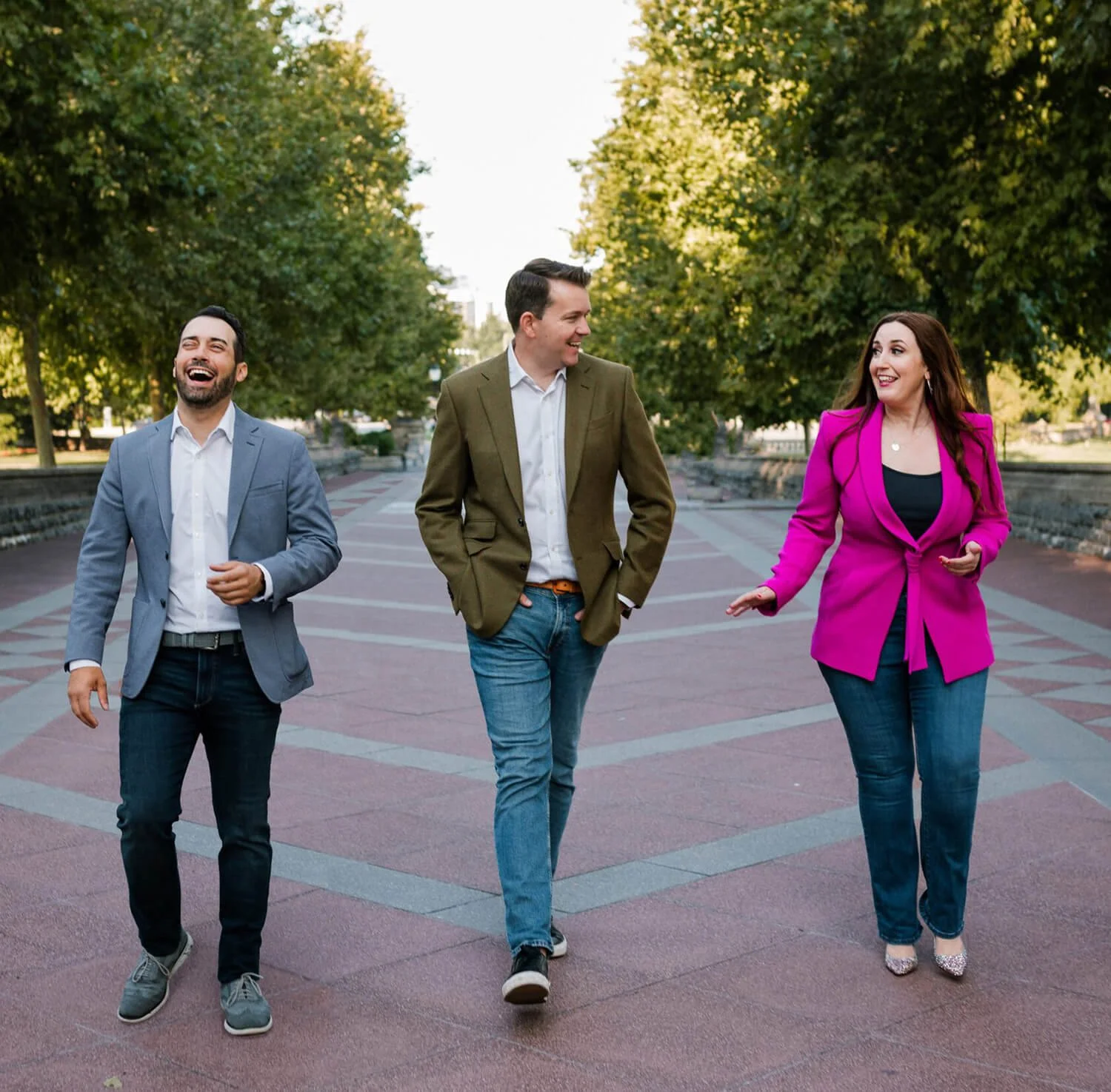 Three friends, two men and one woman, walk and talk in a park with green trees, enjoying a sunny day. They are smiling and engaged in conversation.