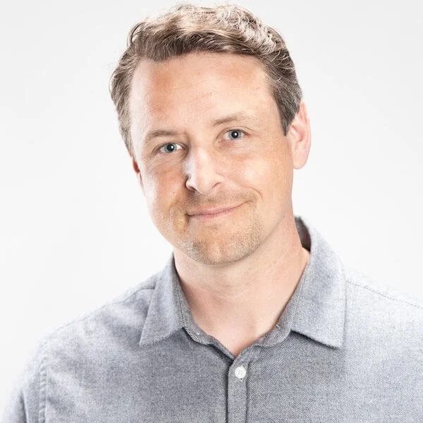 Portrait of a man with light brown hair, blue eyes, and fair complexion, wearing a light gray button-up shirt, smiling slightly, against a plain light background.