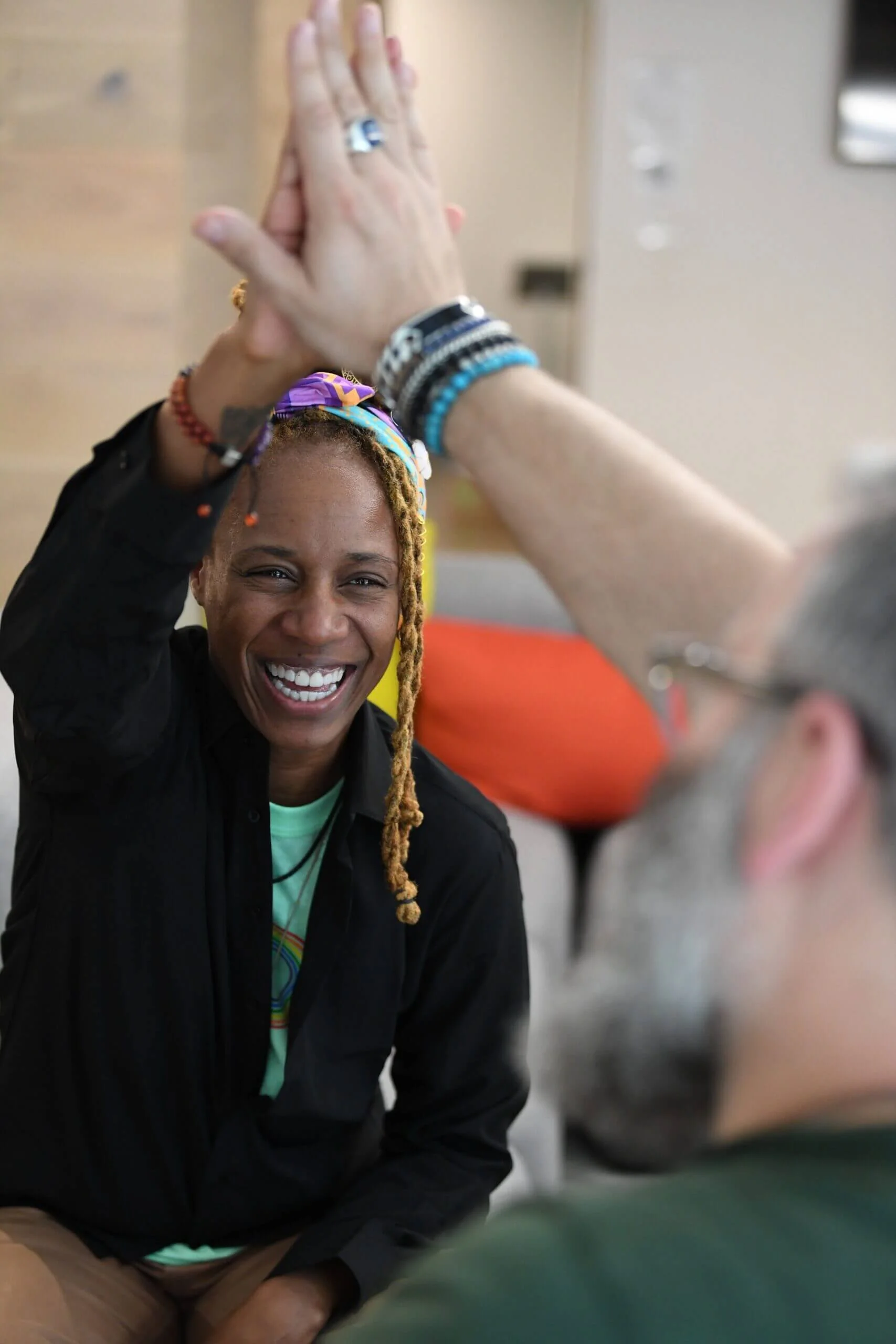 A joyful lack woman with dreadlocks, wearing a colorful headband and a rainbow T-shirt, smiling widely while giving a high five. She is seated indoors with a blurred background and a person with a tattooed arm and multicolored bracelets reaching out to high five her.