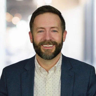 Headshot of a man with a beard, smiling, wearing a dark blazer and a white patterned shirt, in an indoor setting with blurred background.