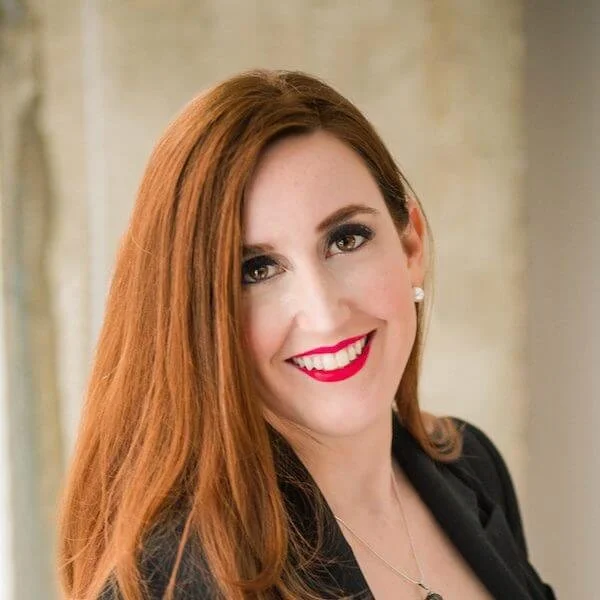 A woman with long red hair and fair skin, smiling with red lipstick, wearing earrings and a black blazer, in front of a light-colored wall.