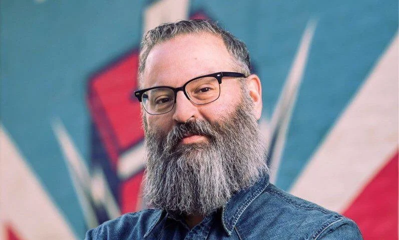 A bearded man wearing glasses and a denim shirt standing in front of a colorful, abstract background.