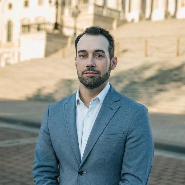 A man in a blue suit and white shirt standing outdoors with steps and historic buildings in the background