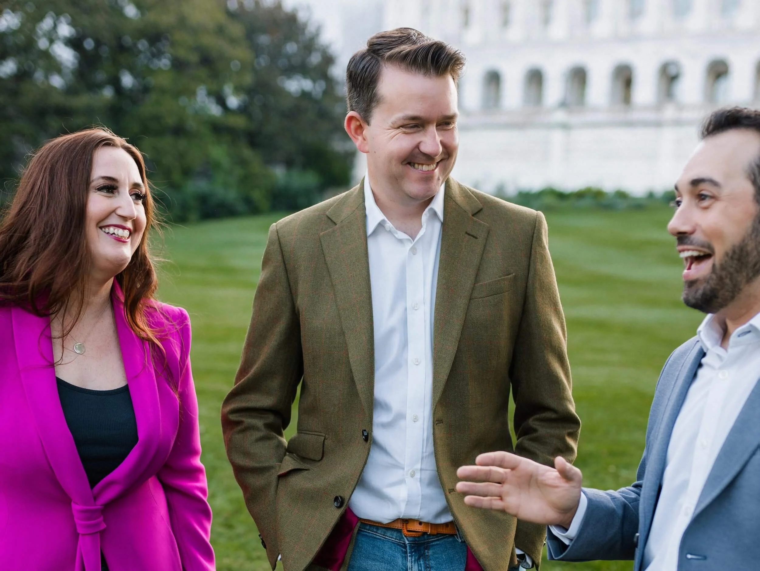A group of people, smiling and talking outdoors on a green lawn with trees and a white building in the background.
