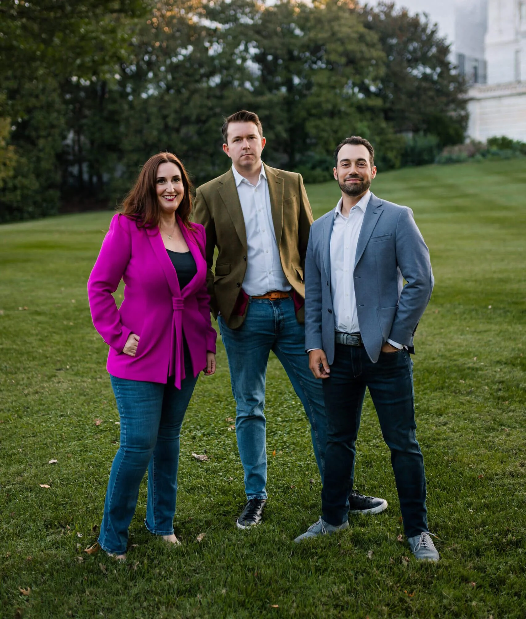 Three professionally dressed people standing outdoors on a grassy area with trees and a white building in the background.