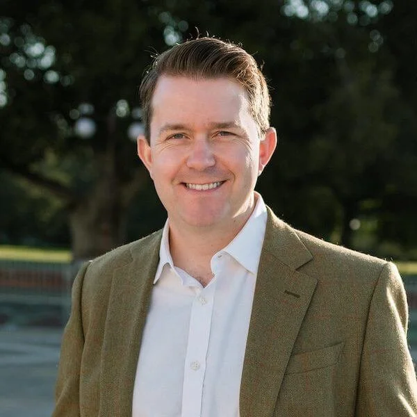 A man wearing a beige blazer and white shirt, smiling outdoors with trees in the background.