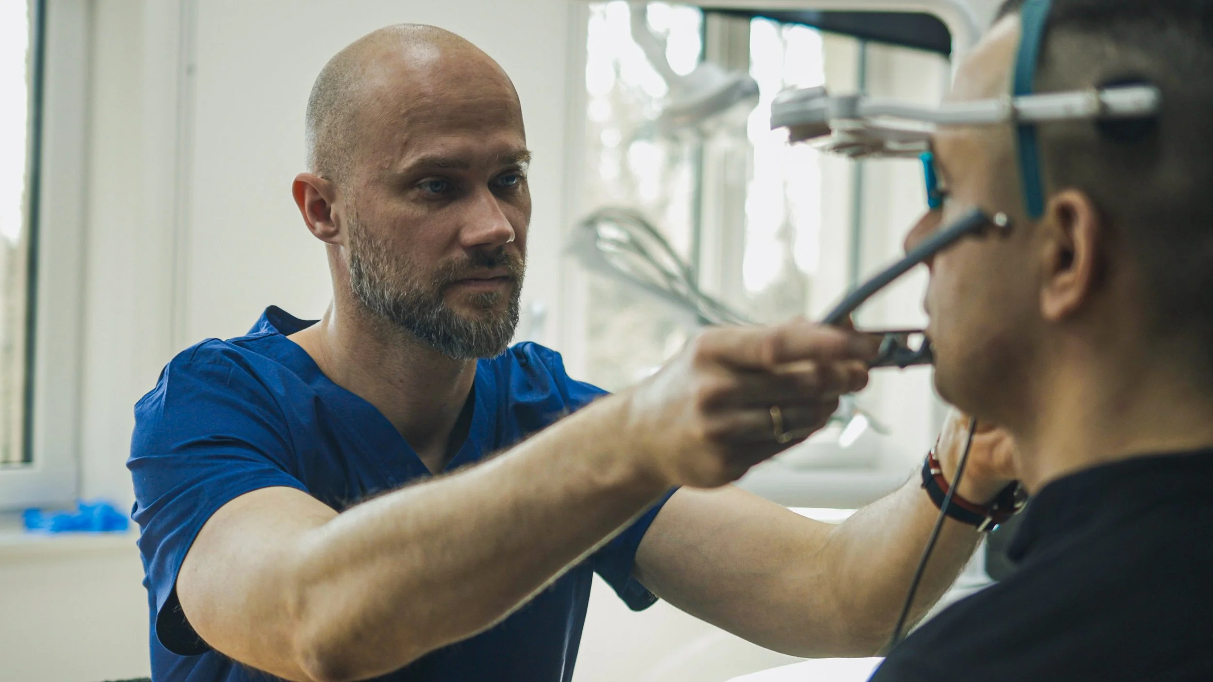 A healthcare professional in blue scrubs examining a patient's ear with specialized medical equipment in a clinical setting.