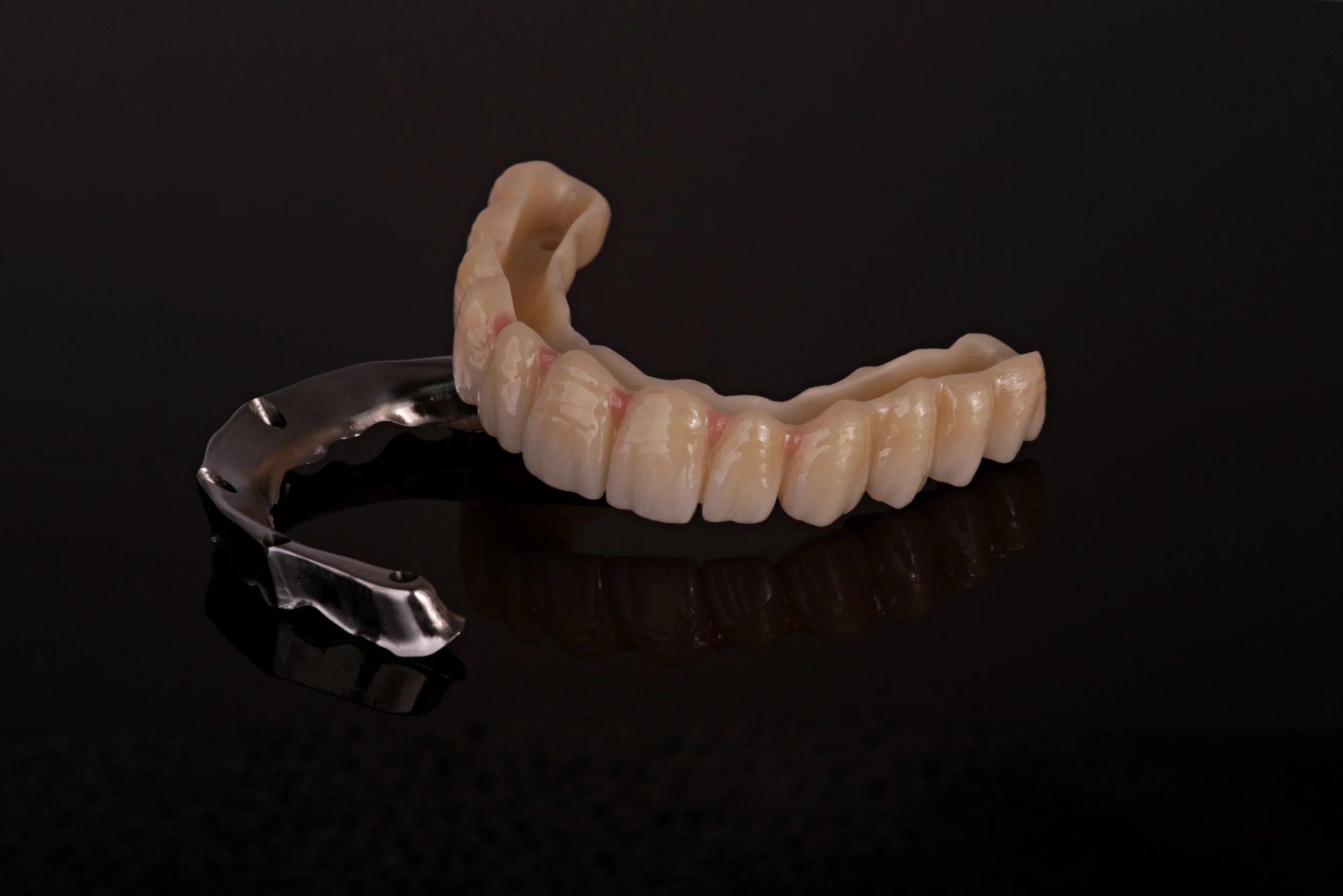 A set of dental dentures, containing upper and lower artificial teeth, placed on a reflective black surface with a dark background.
