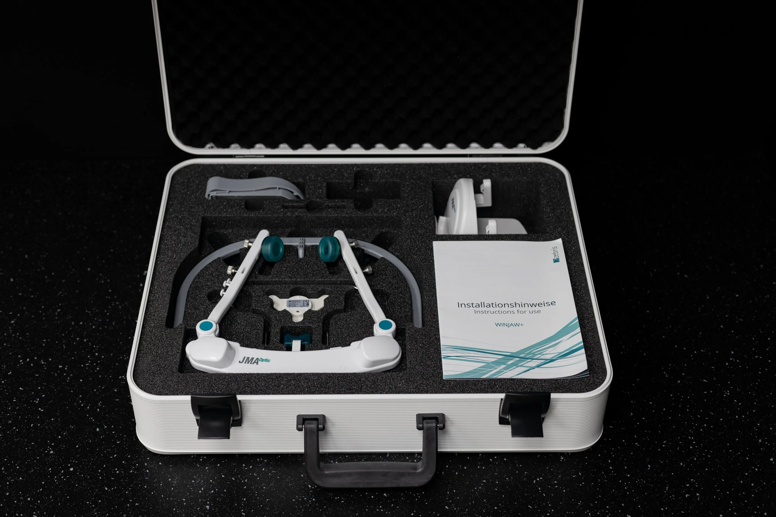 Open white hard case containing eye tracking device with foam insert, instruction manual, and accessories on a black speckled surface.