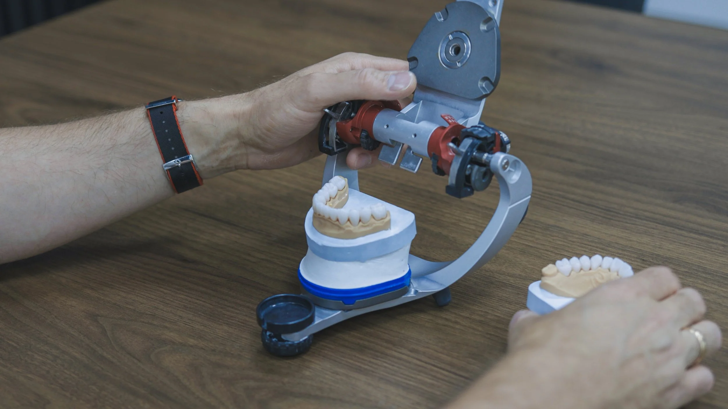 A person wearing a black watch with red stitching working with a dental or jaw model on a wooden table, using a specialized mechanical device.