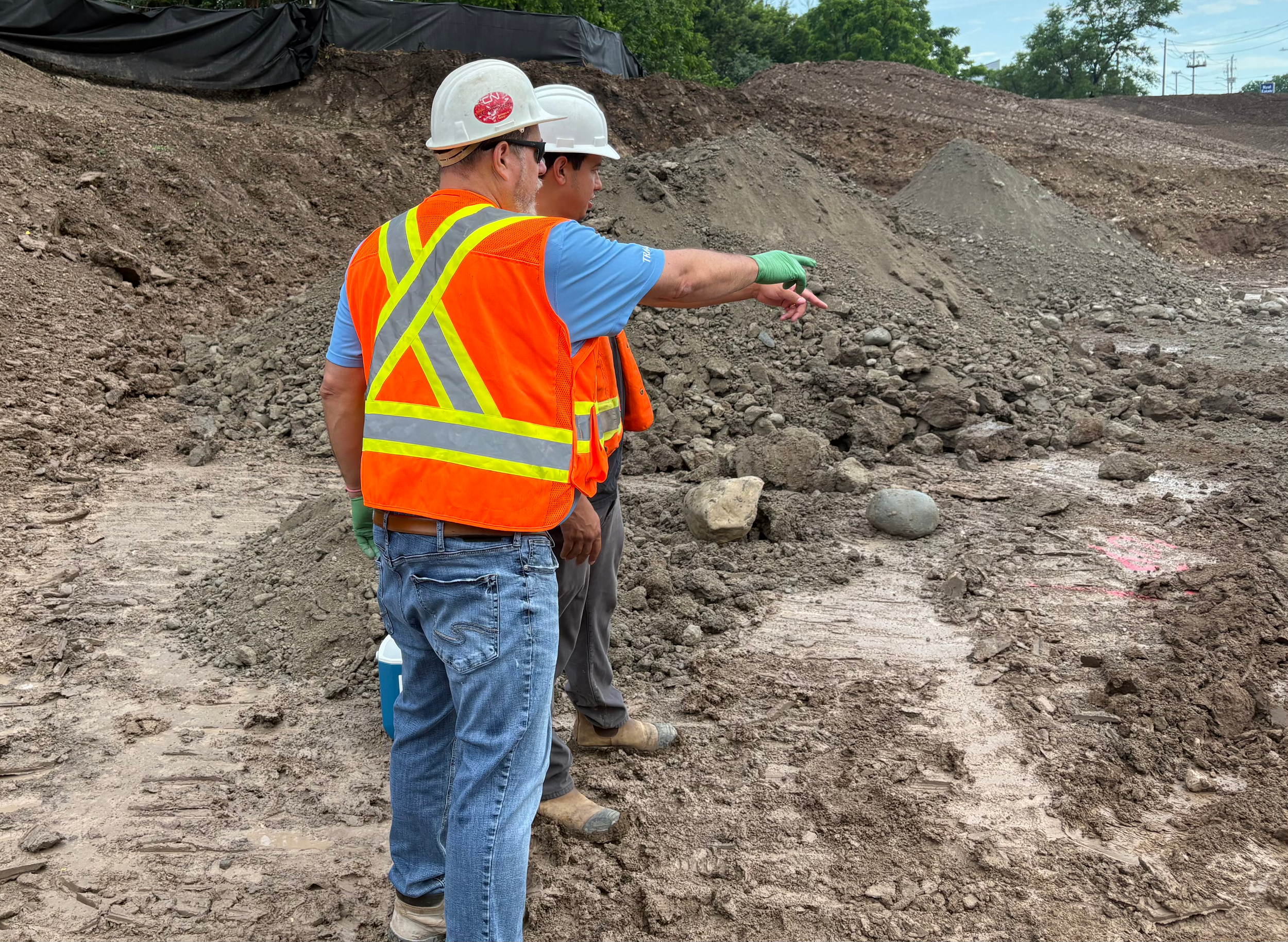 Two construction workers, wearing white helmets and reflective vests, stand outdoors on a construction site, inspecting a pile of dirt and rocks while pointing at something in the distance.