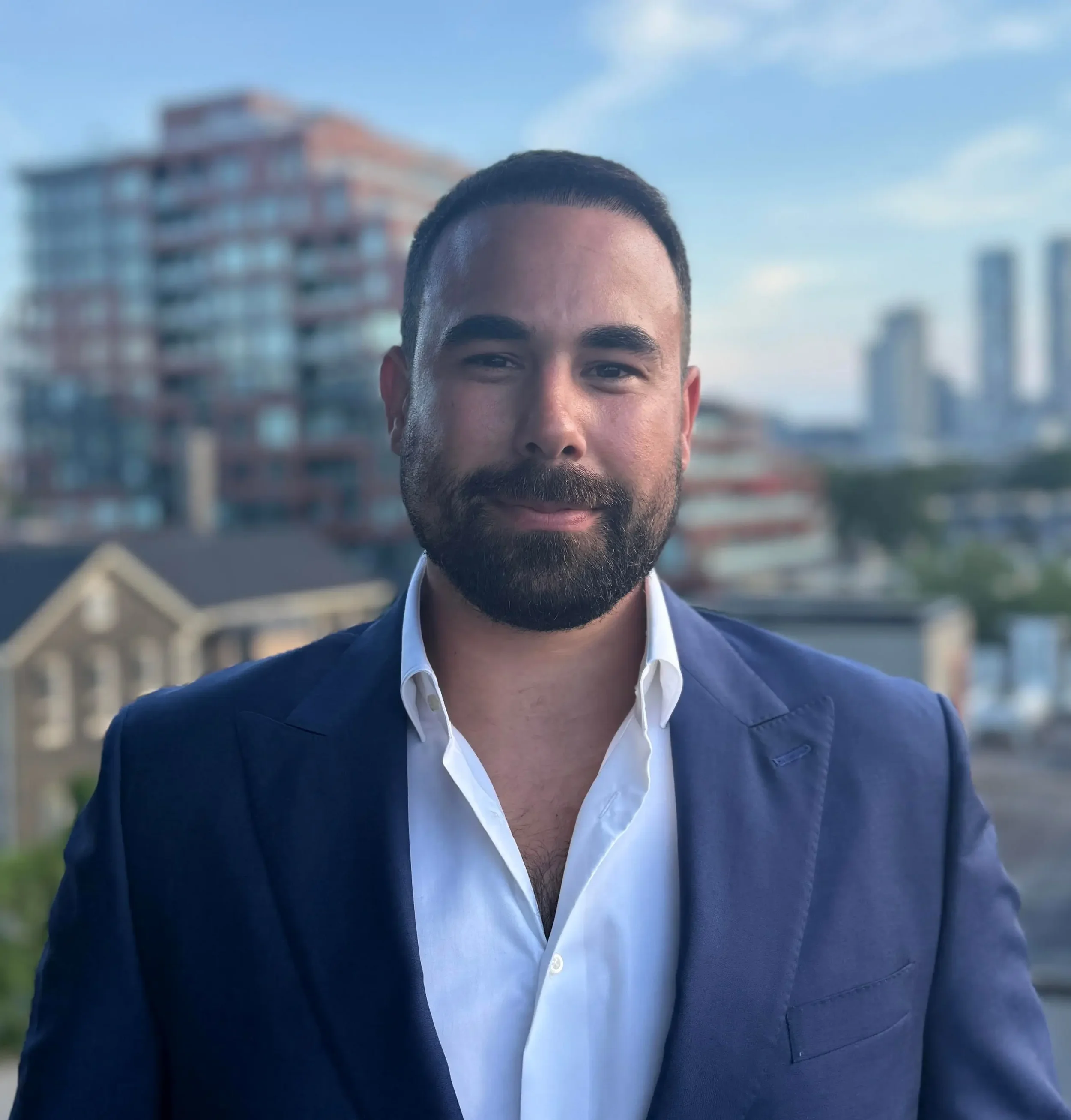 Close-up of a man with a beard and short dark hair, wearing a navy blazer and a white dress shirt, standing outdoors with a cityscape of tall buildings behind him under a partly cloudy sky.