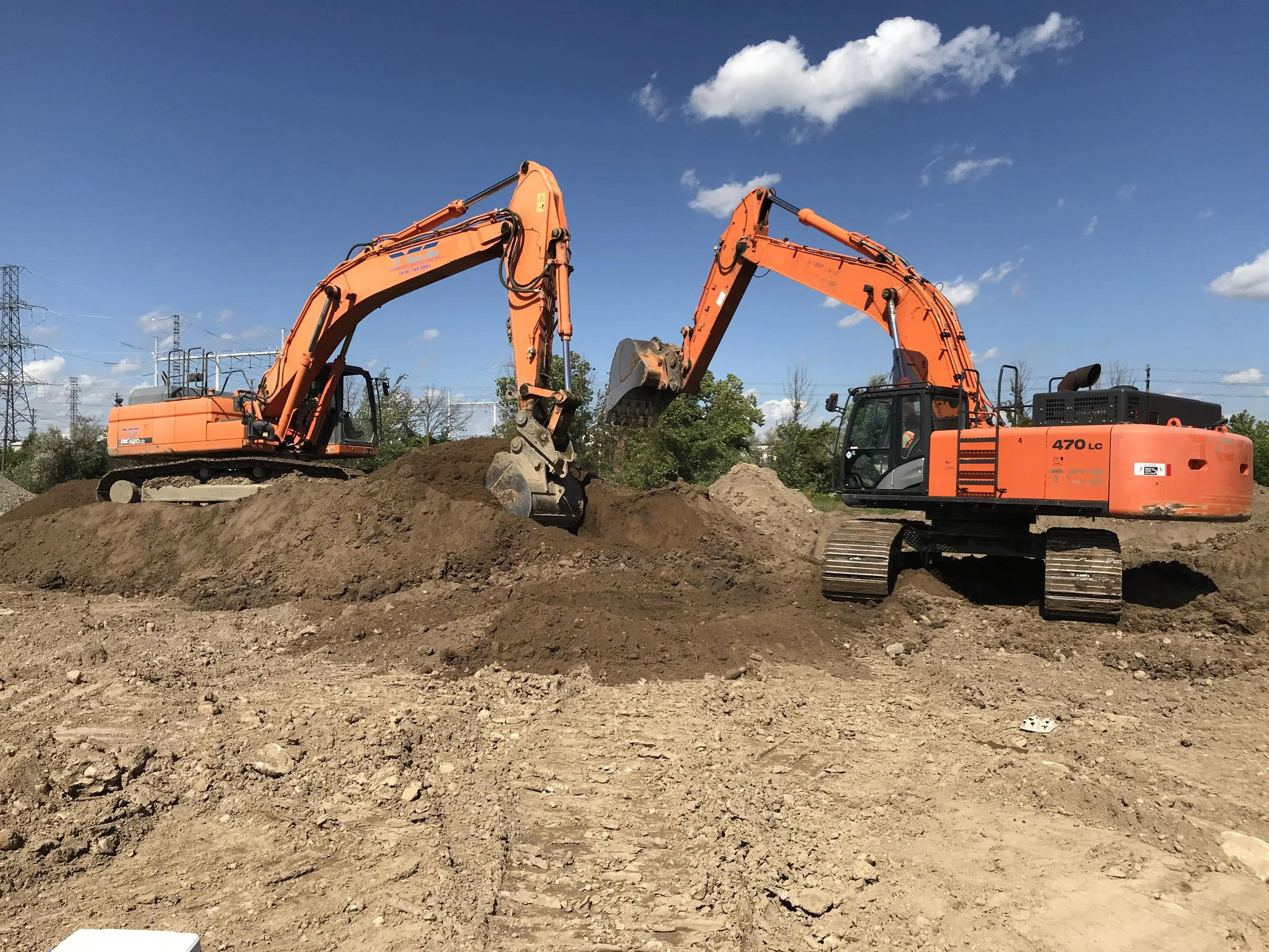 Two orange excavators are working on a construction site with dirt and soil, under a blue sky with some clouds, and power lines in the background.