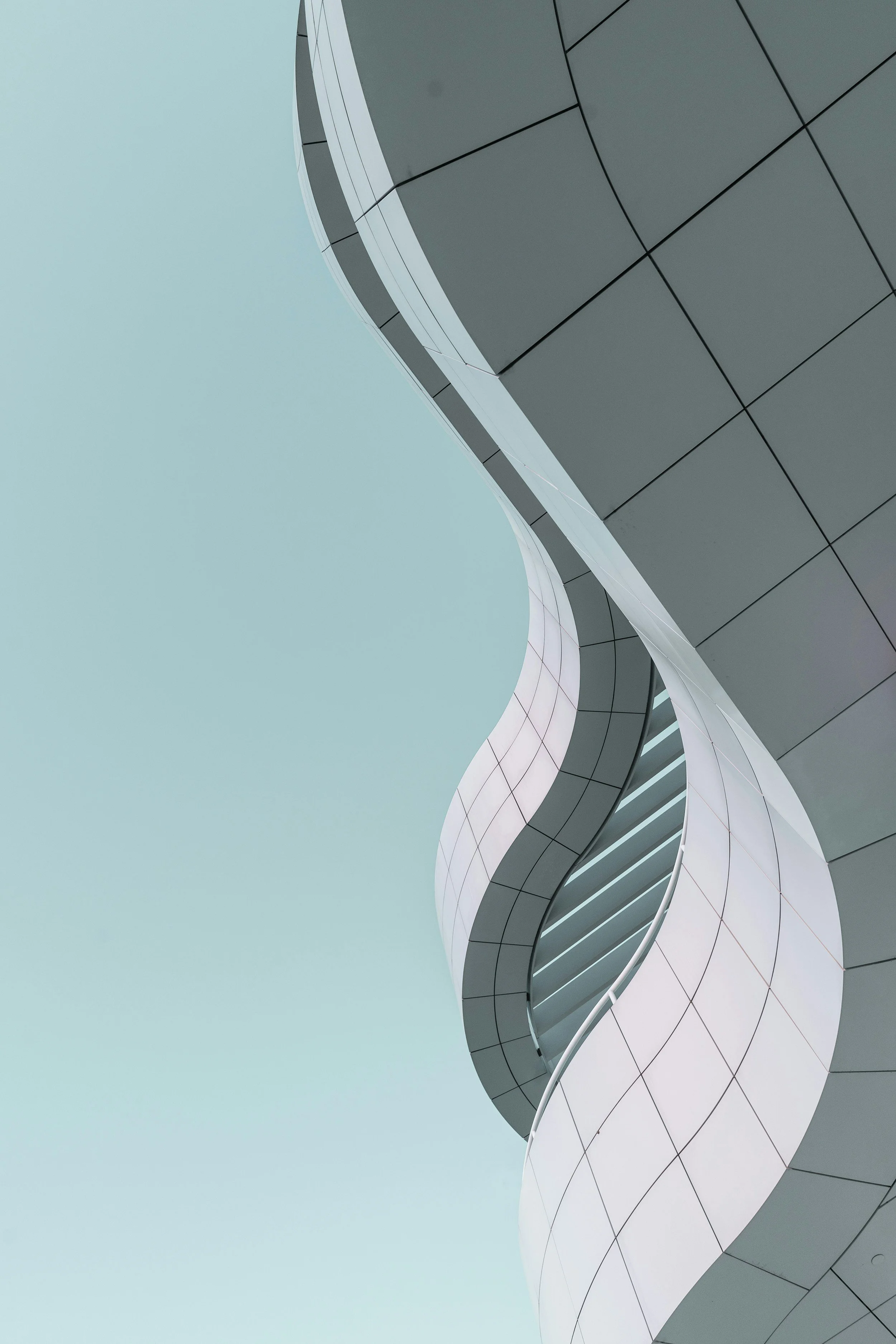 Close-up view of a modern architectural building with curved lines, gray panels, and white accents, against a pale blue sky.