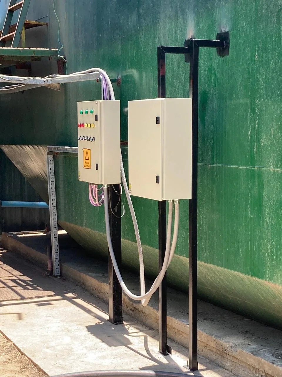 Panel eléctrico y cables instalados en una estructura metálica cerca de un tanque grande de color verde.