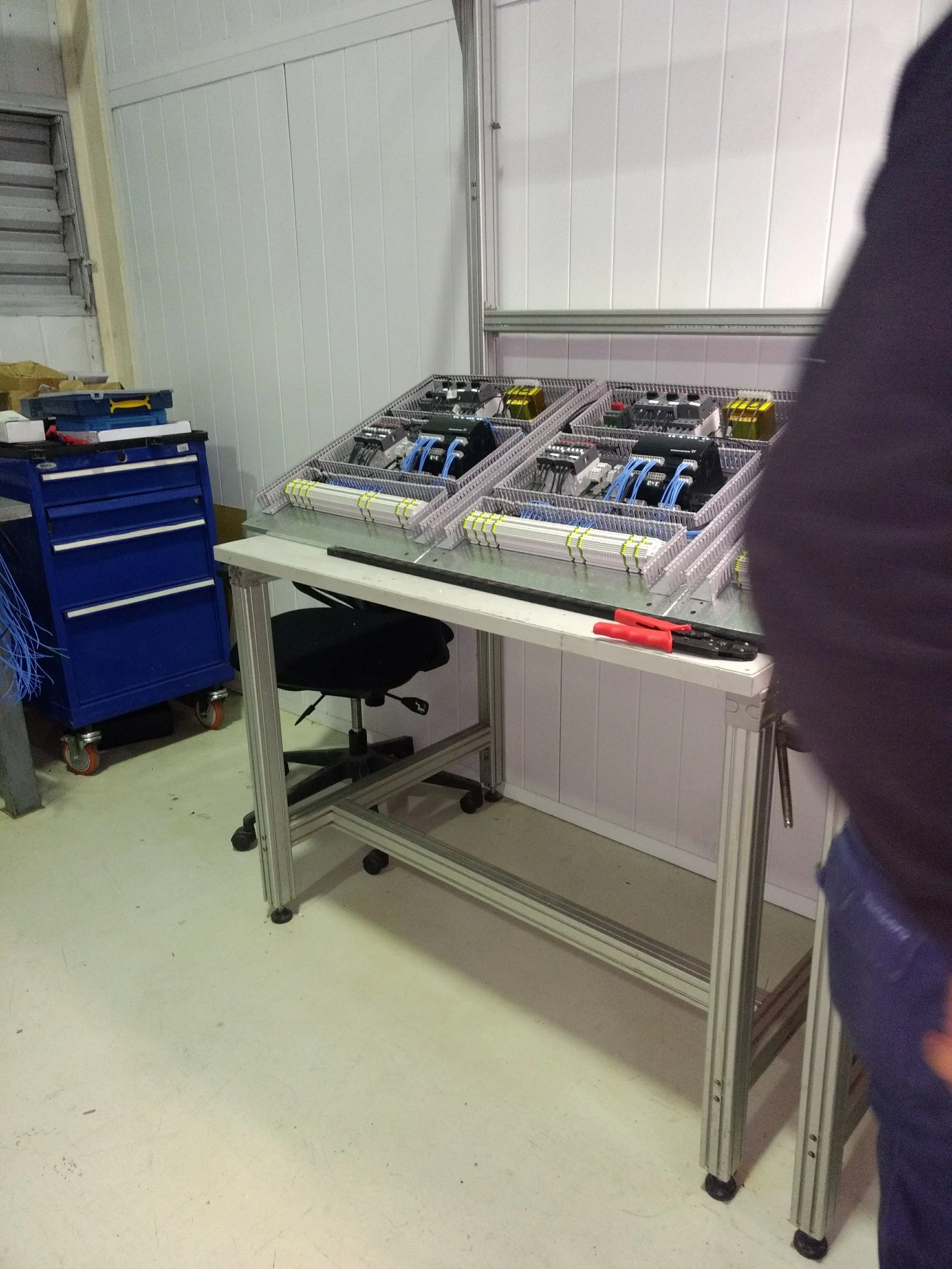 Mesa de trabajo con componentes electrónicos en cajas de separación, herramientas y una silla, en una sala técnica con paredes blancas y piso claro.