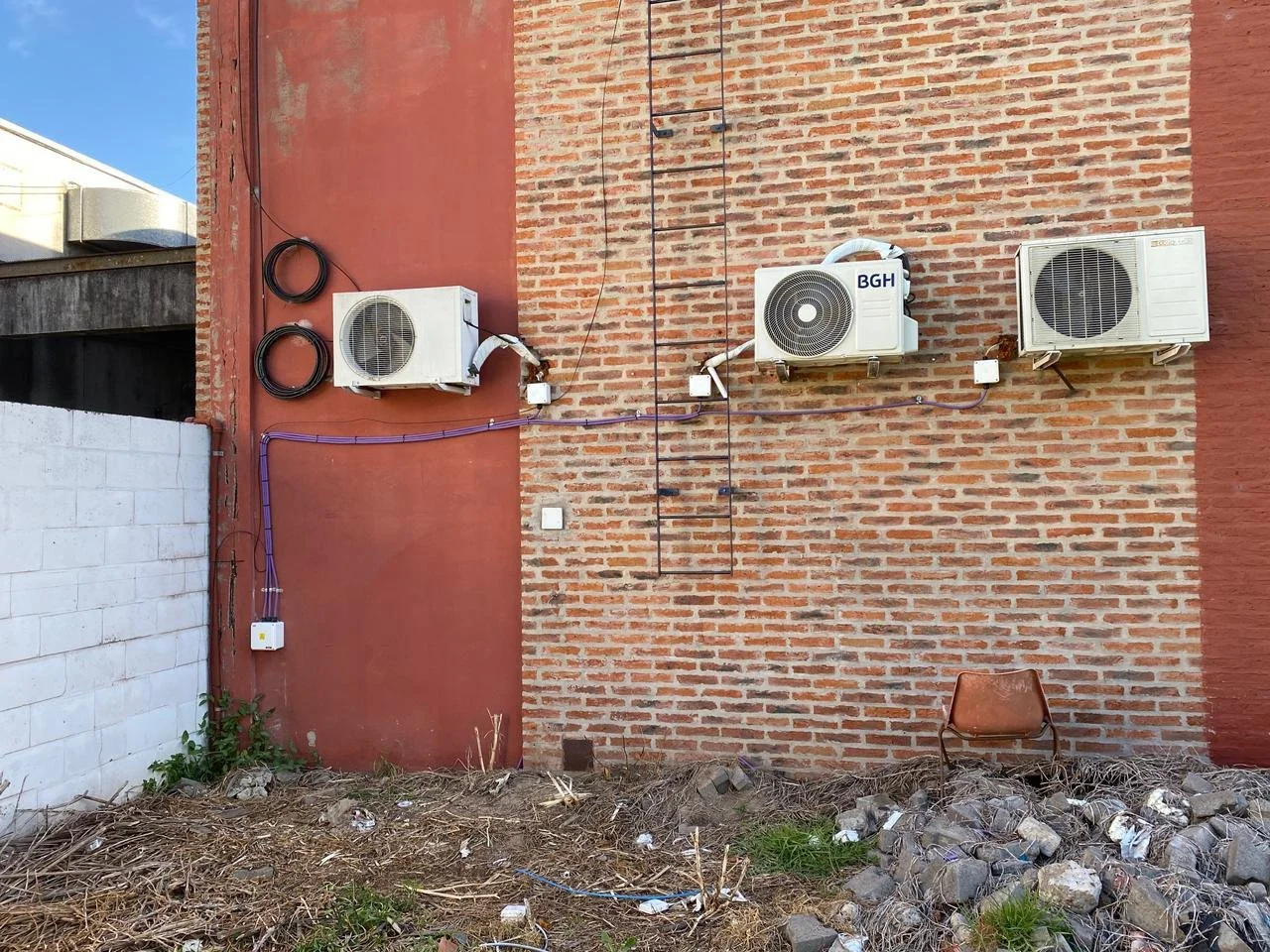 Pared de ladrillo con tres unidades de aire acondicionado y cables de electricidad, un mueble viejo y basura en el suelo.