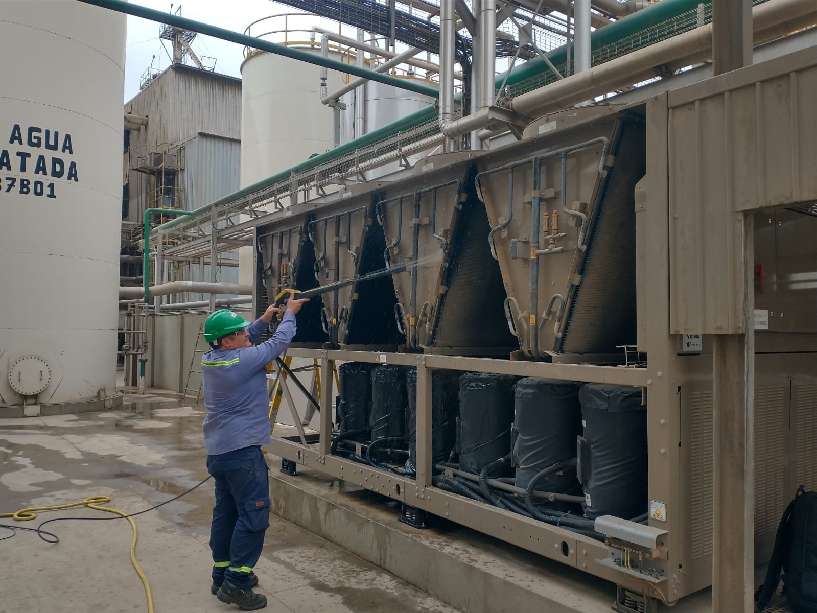 Trabajador con casco verde limpiando un equipo industrial en una planta de agua tratada con una manguera.