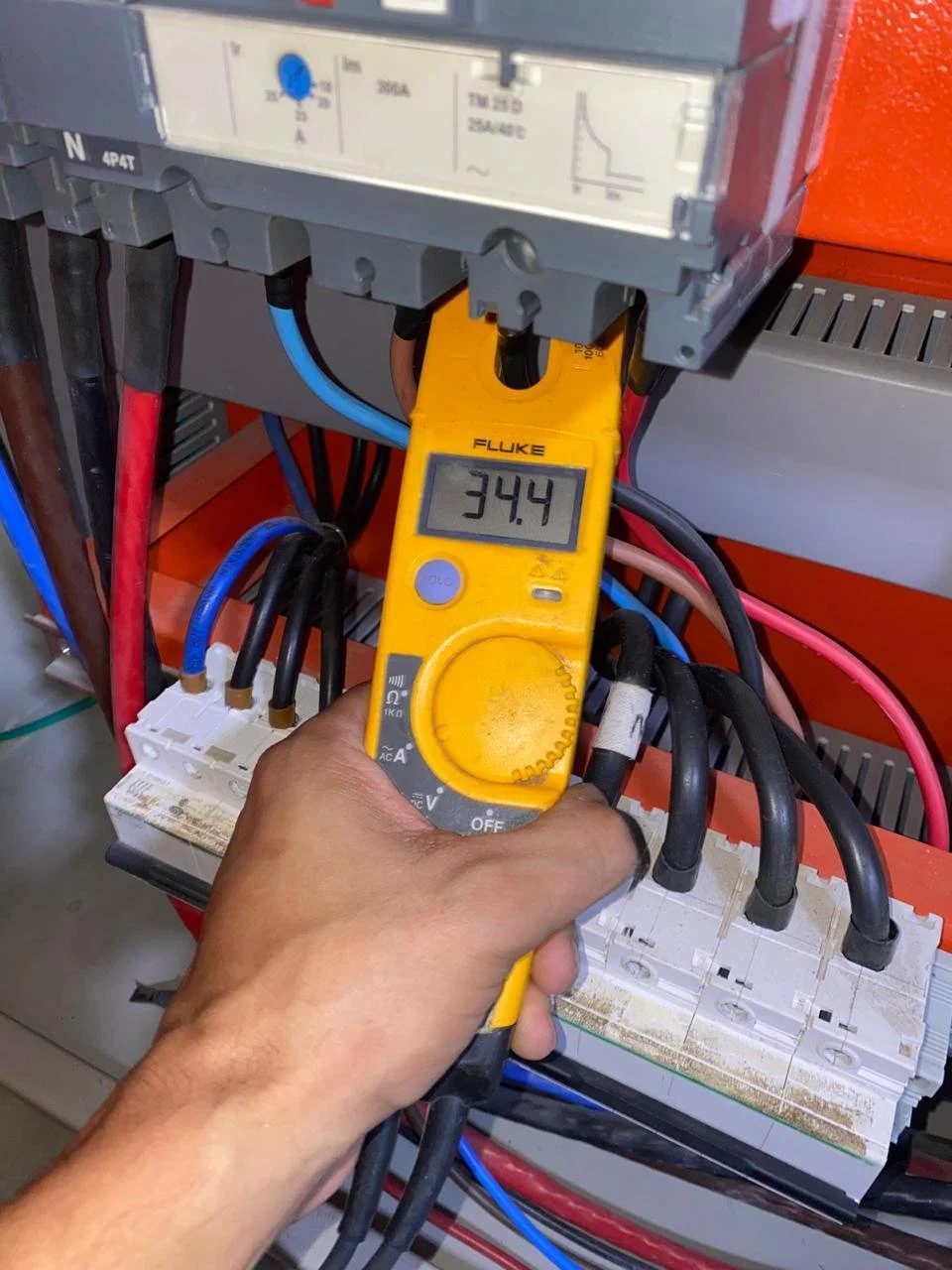 Persona usando un multímetro digital amarillo para medir 34.4 voltios en un panel de control eléctrico con cables de diferentes colores y componentes electrónicos.