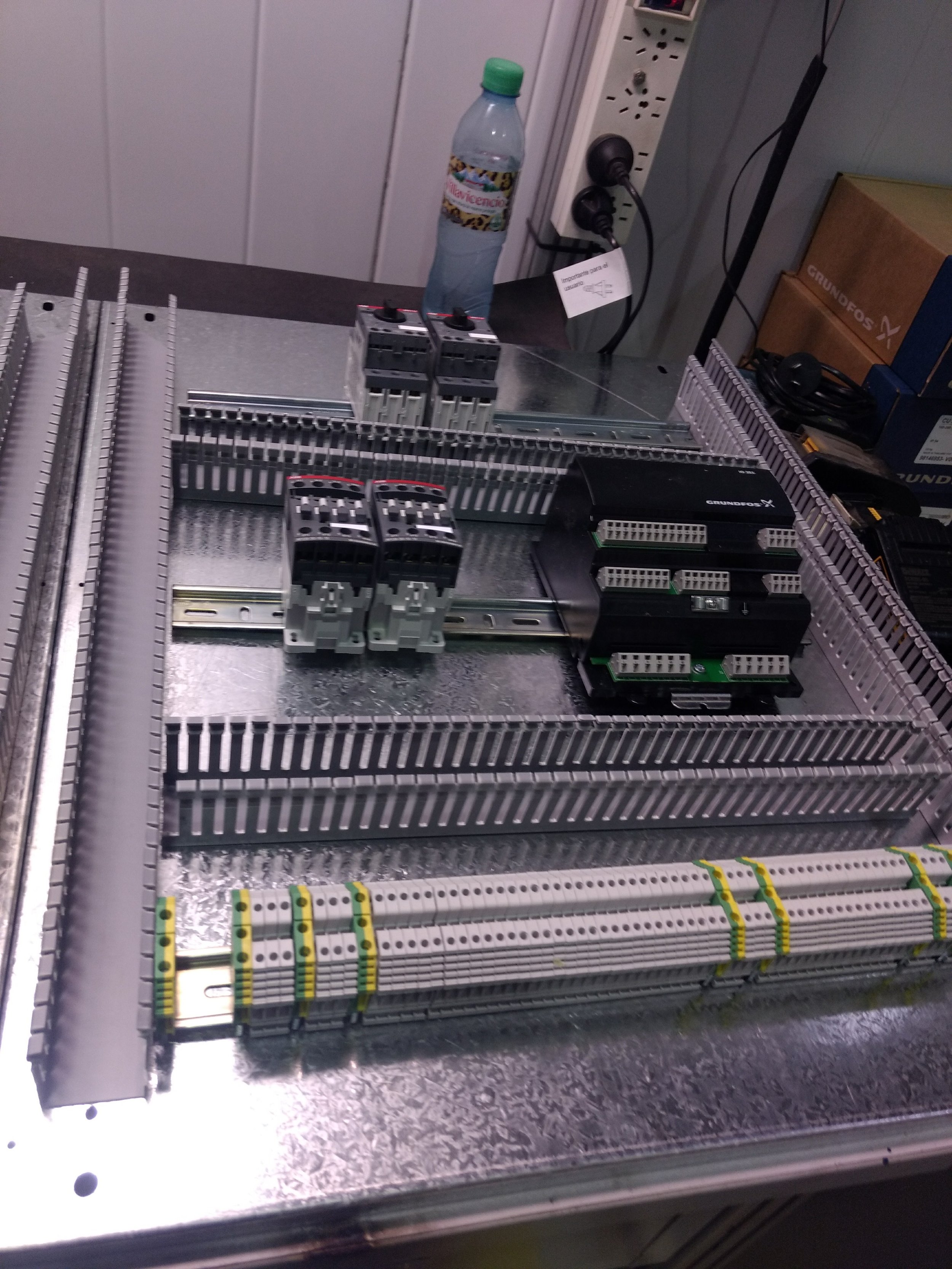 Tableros de control eléctrico con componentes electrónicos y conexiones, una botella de agua y un enchufe de corriente en un área industrial o de maquinaria.