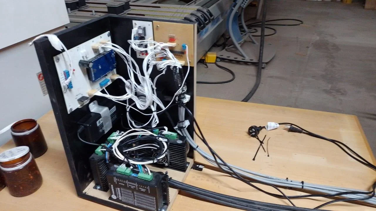 Componentes electrónicos y cables en una carcasa de metal sobre una mesa de madera, con cables conectados y una toma de corriente en la parte derecha.
