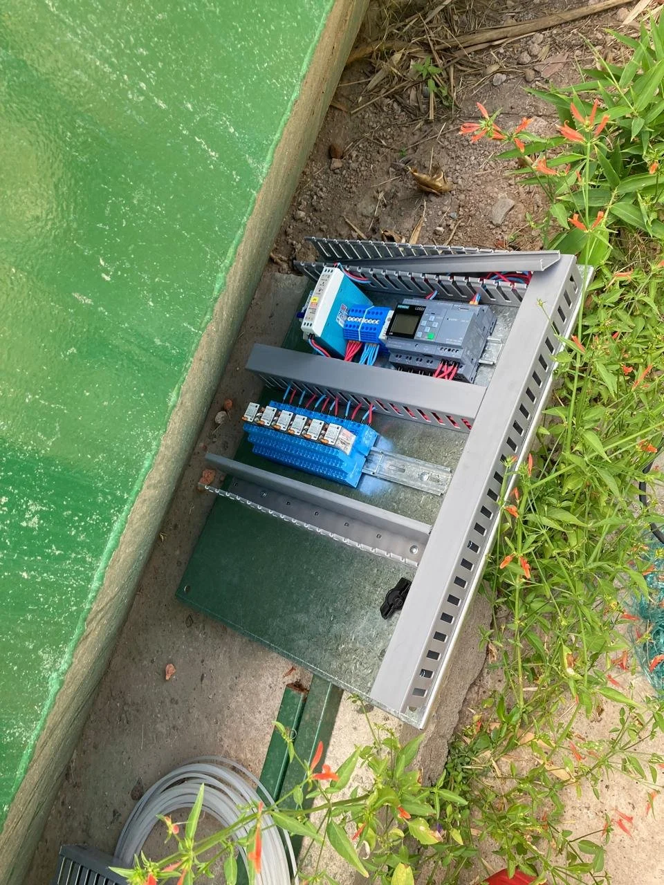 Panel de control y componentes eléctricos instalados en el suelo, rodeados de plantas y cerca de una pared verde.