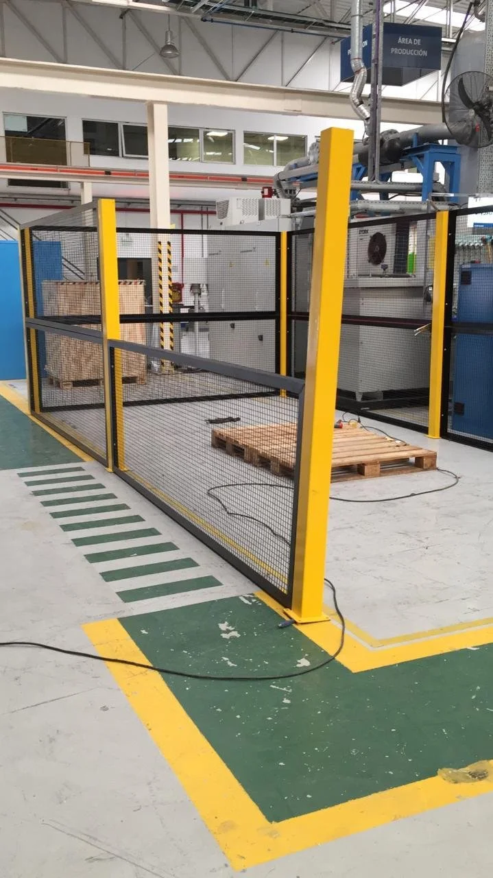 Zona de seguridad con valla metálica en una fábrica, cerca de maquinaria industrial y pallets de madera, en un área de producción.