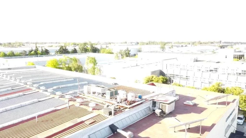 Vista de techo de un edificio con equipo de aire acondicionado y estructuras sobre él, con fondo de plantas y otros edificios.