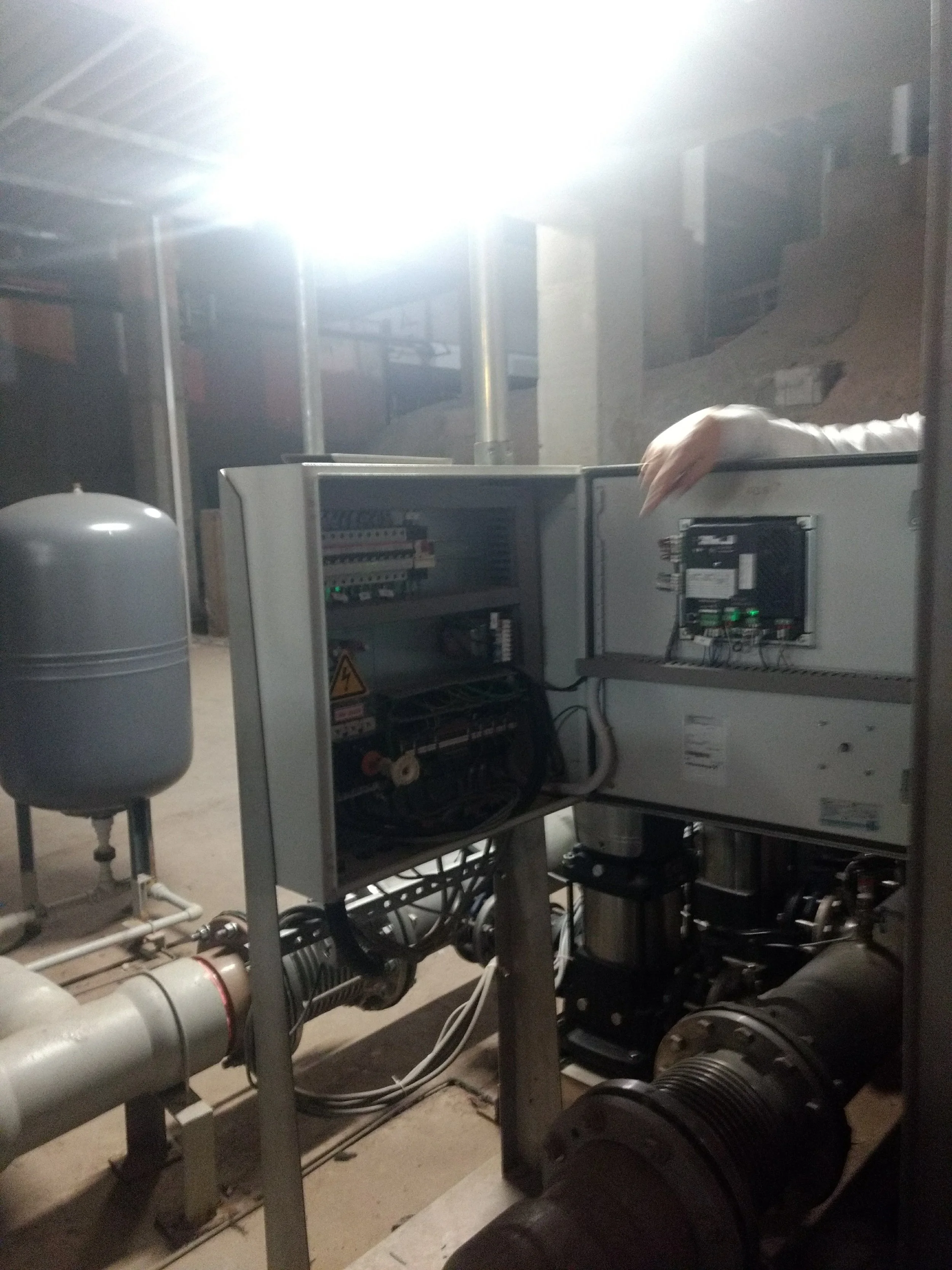 Cuadro de control eléctrico con componentes y cables en un entorno industrial, con tuberías y mecanismos visibles alrededor.