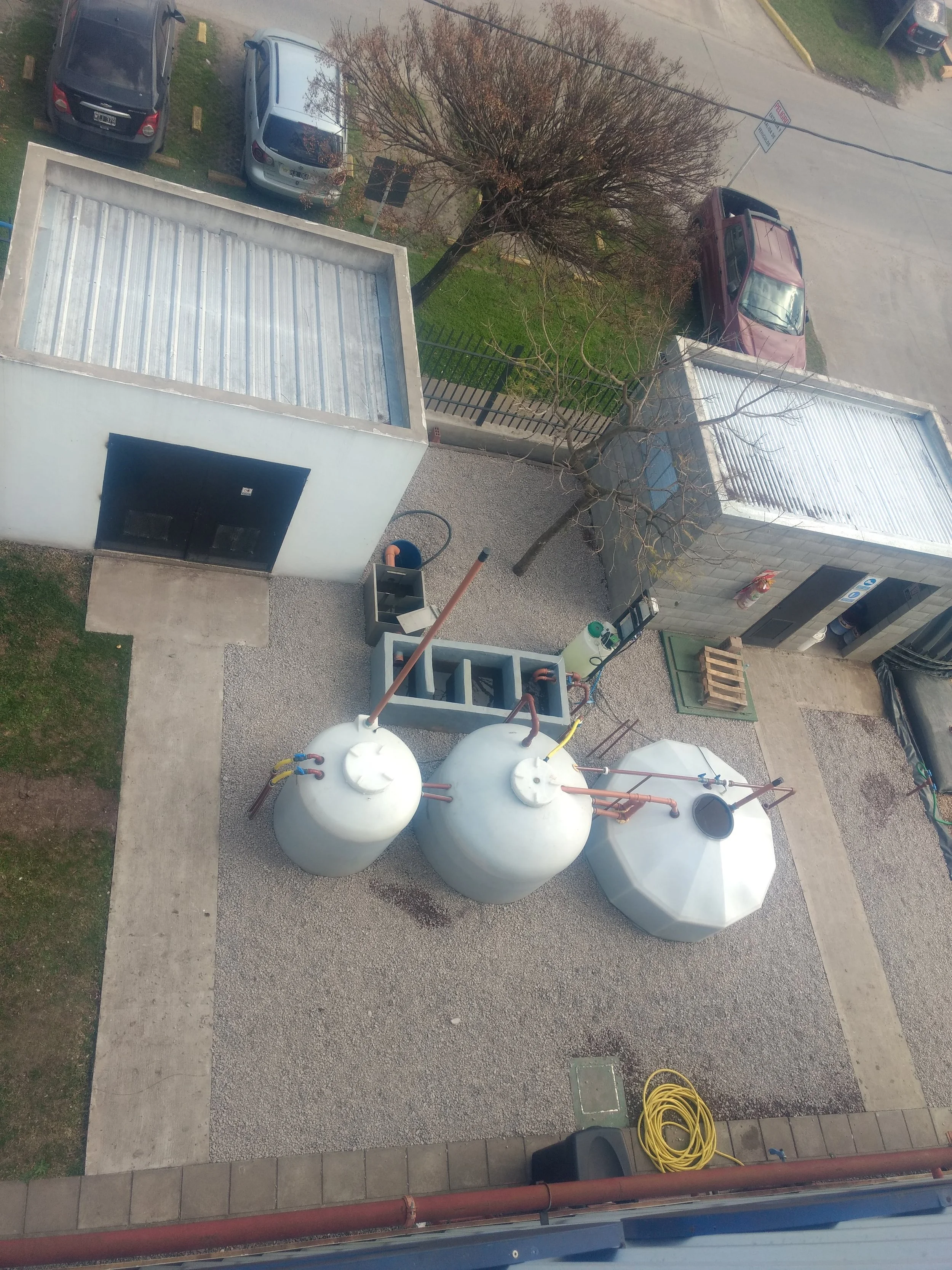 Vista de un área exterior con tanques de almacenamiento, herramientas y vehículos estacionados. Incluye un árbol sin hojas, un cable de electricidad, un garaje y una calle con más automóviles.