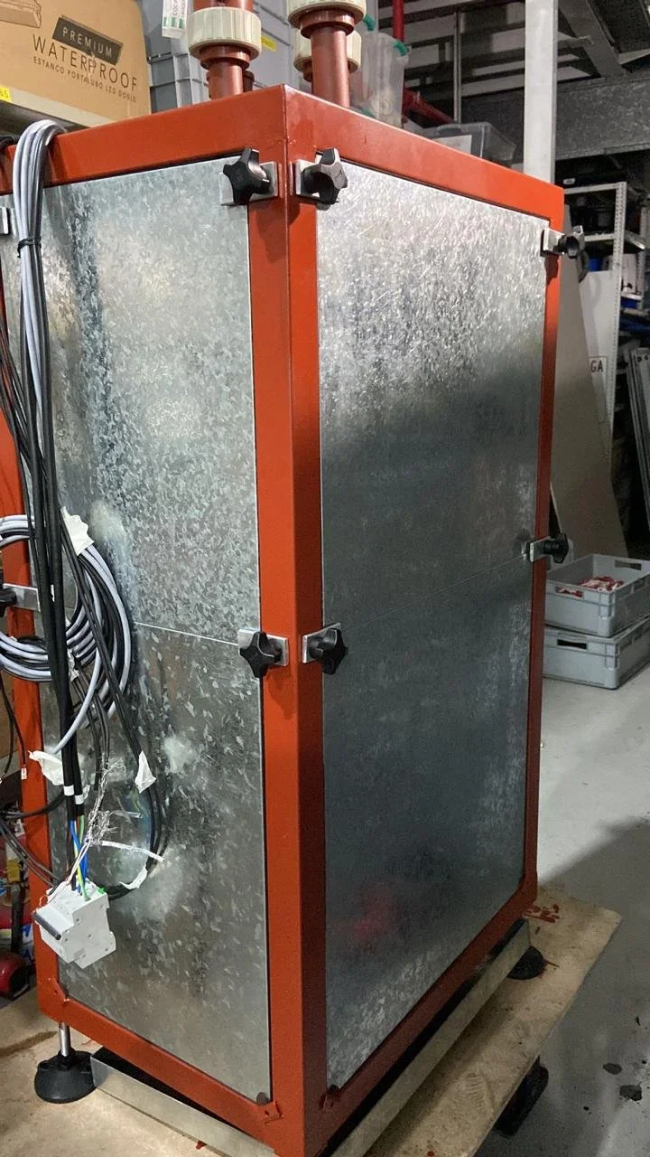 Equipamiento industrial con estructura metálica naranja y paredes de metal, ubicado en un taller con cables y herramientas en el fondo.