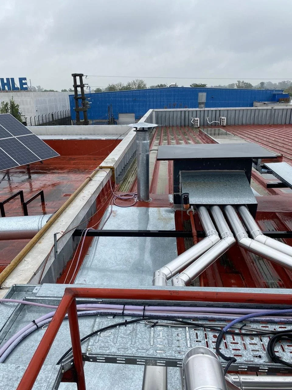 Techo de una azotea con paneles solares, tuberías de ventilación, cables y estructuras metálicas, con cielo nublado y edificios azules y blancos en el fondo.