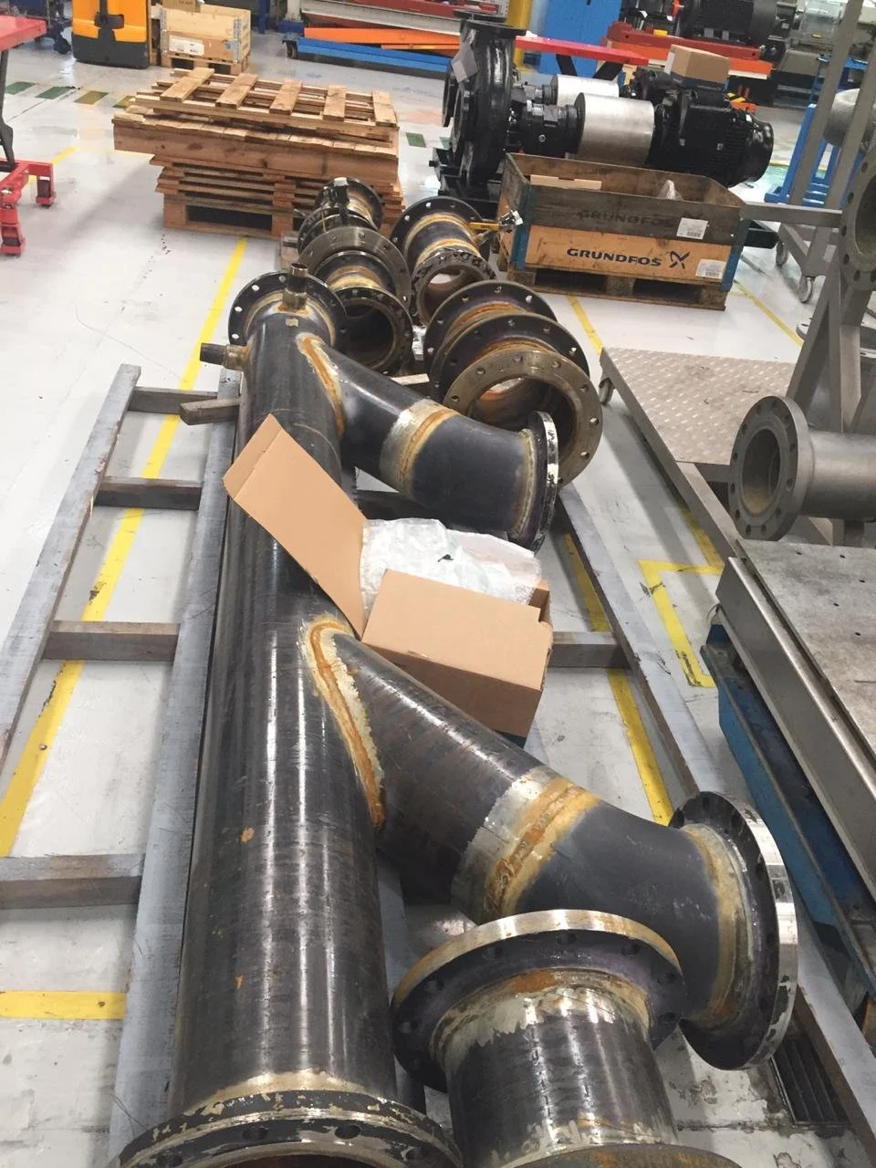 Tubería grande de metal en una fábrica, con varias juntas y partes, área de trabajo con cajas de madera y maquinaria.