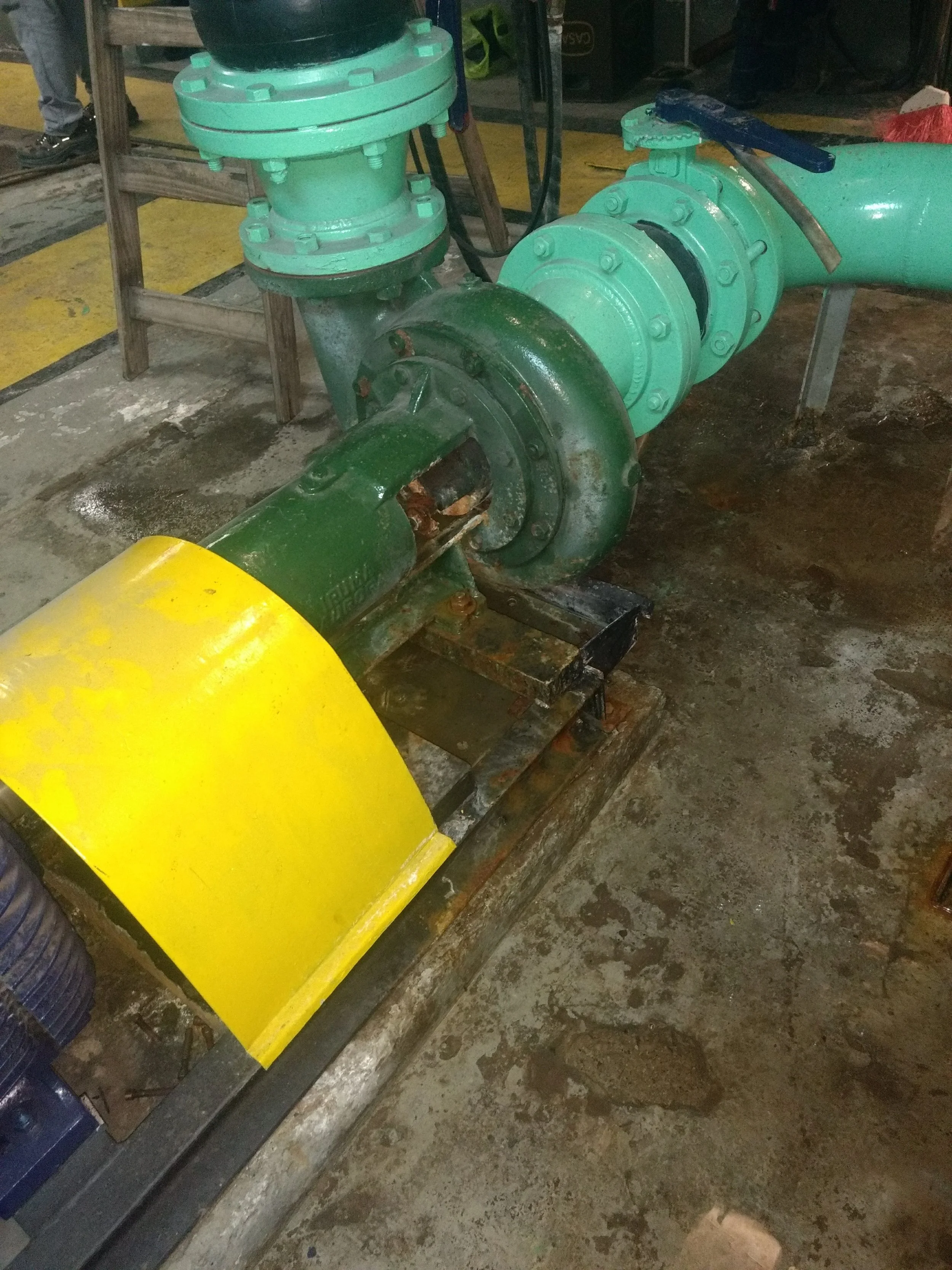 Tubería industrial de color verde con válvula en un entorno de fábrica o planta de producción, sobre piso de concreto. Una parte de un vehículo amarillo está visible en la esquina inferior izquierda.