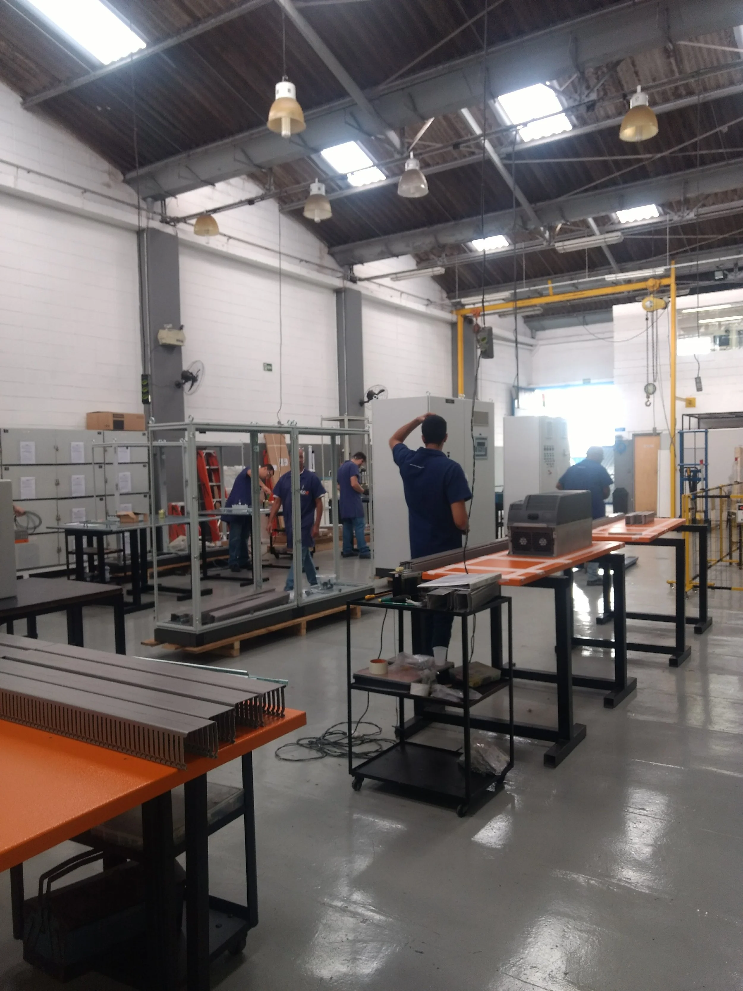 Interior de una fábrica con varias personas trabajando, mesas con componentes electrónicos y maquinaria industrial.