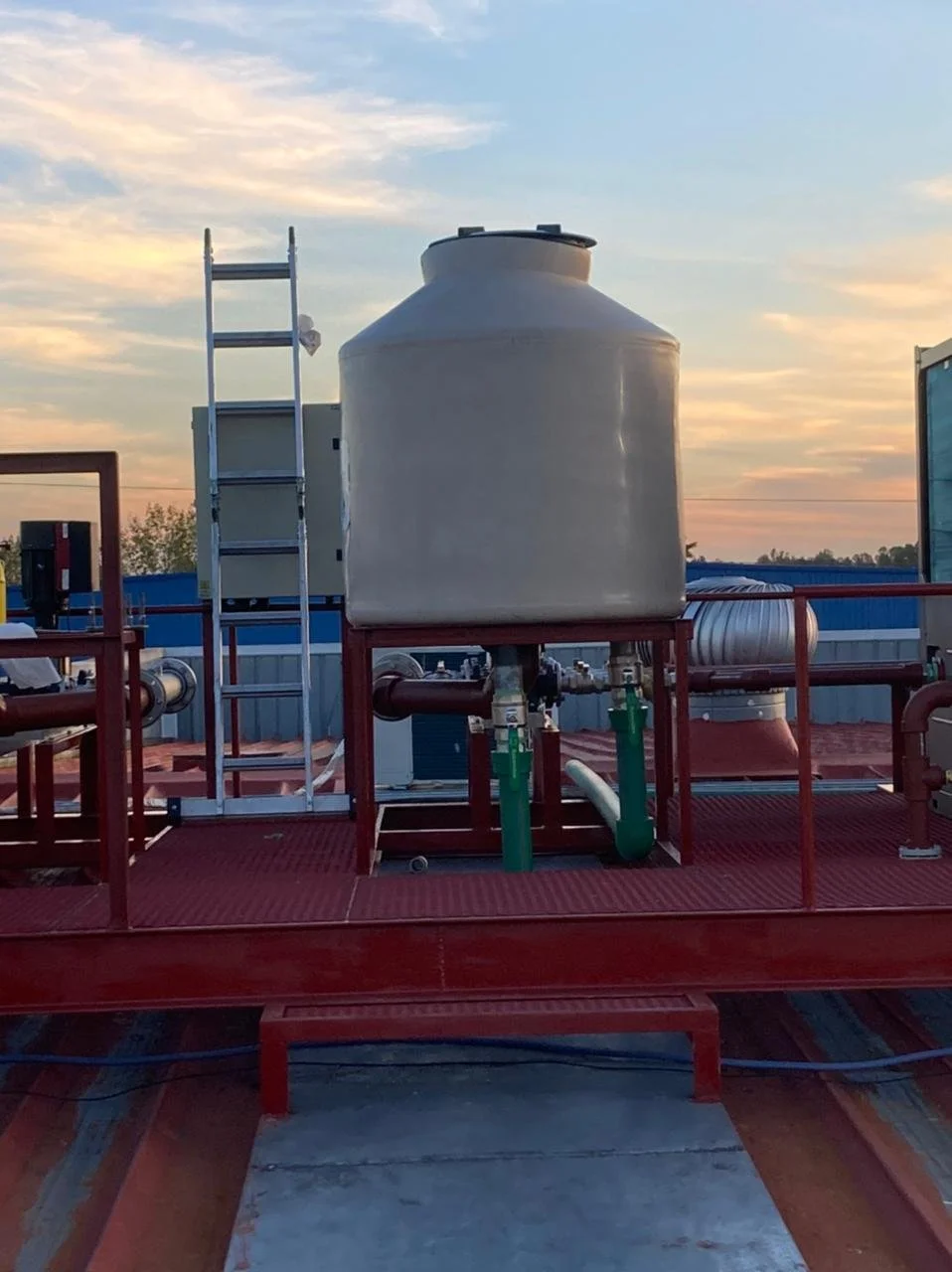 Tanque grande en una plataforma con tuberías verdes, una escalera metálica y un fondo de cielo con nubes y atardecer.