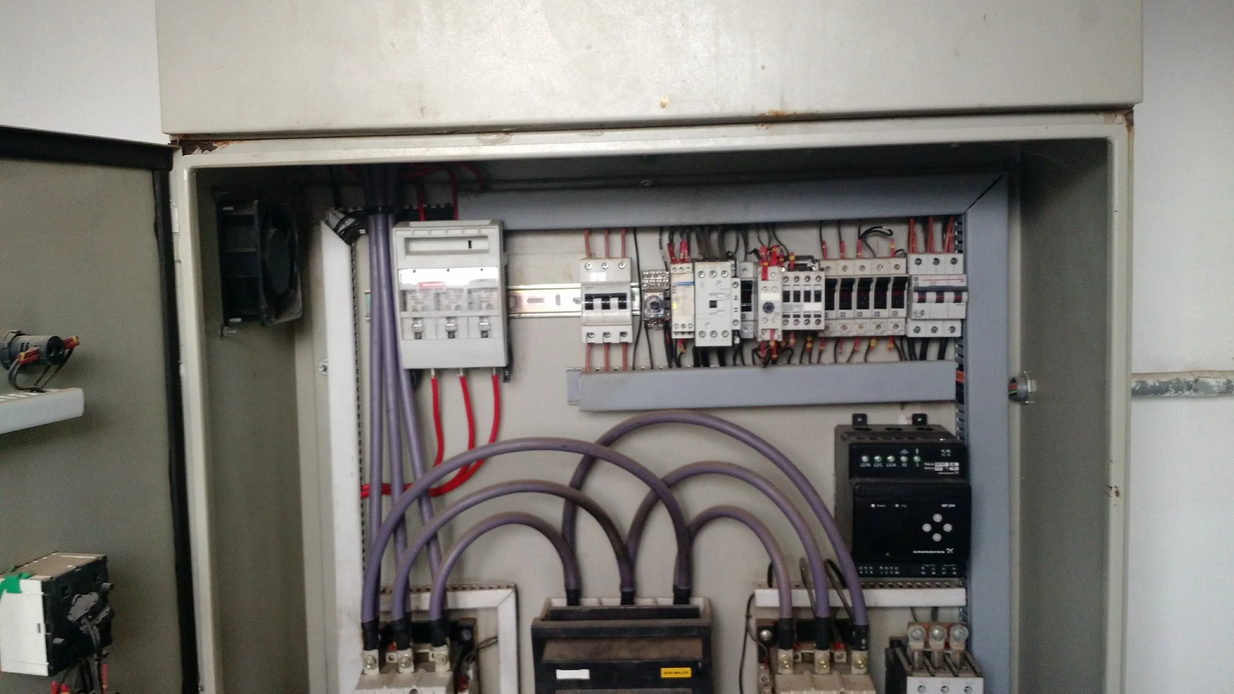 Cuadro eléctrico con componentes y cables internos, incluyendo disyuntores, relés y otros dispositivos electrónicos, con algunos cables morados y rojo. Detrás de una estructura de metal.