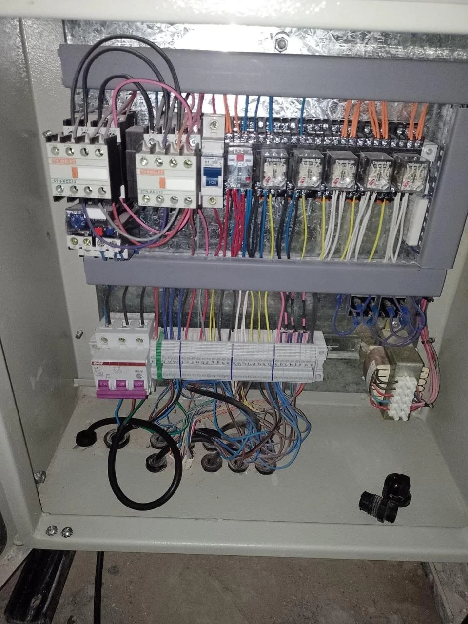 Caja eléctrica con componentes y cables eléctricos internos visible, incluyendo relés, fusibles, y conexiones de cables en diferentes colores.