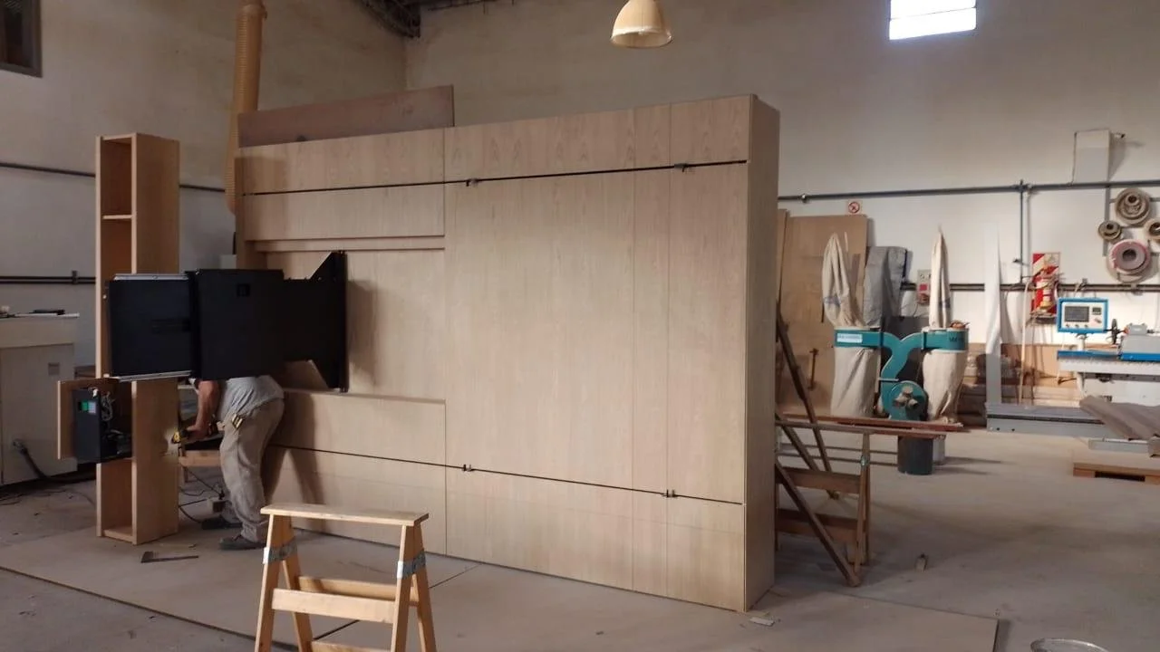 Obra de madera en un taller de carpintería con herramientas y maquinaria, con una estructura de madera en construcción y una persona trabajando.