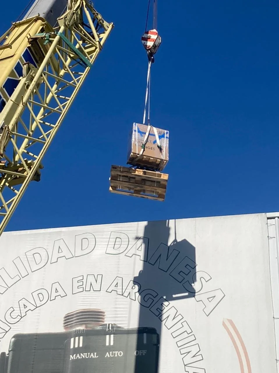 Una grúa de construcción elevando una caja de mercancía y una paleta de madera contra un cielo despejado.
