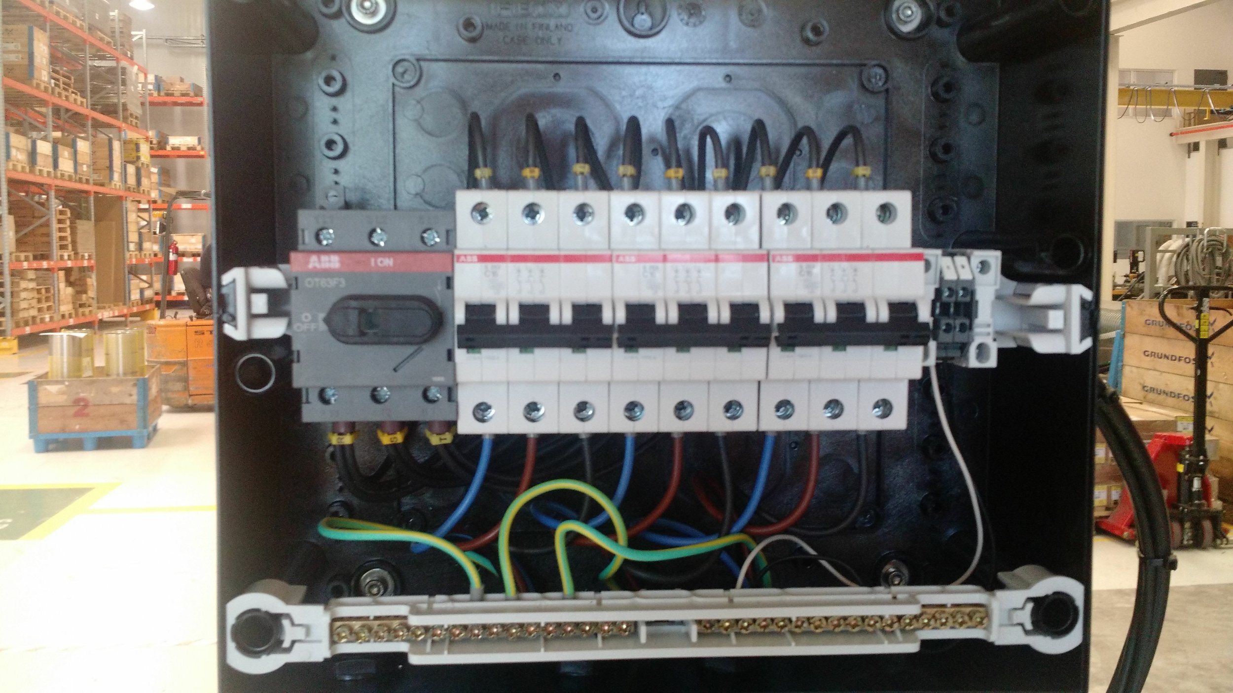 Cuadro eléctrico con componentes electrónicos y cables en un ambiente industrial, con estanterías y cajas visibles en el fondo.