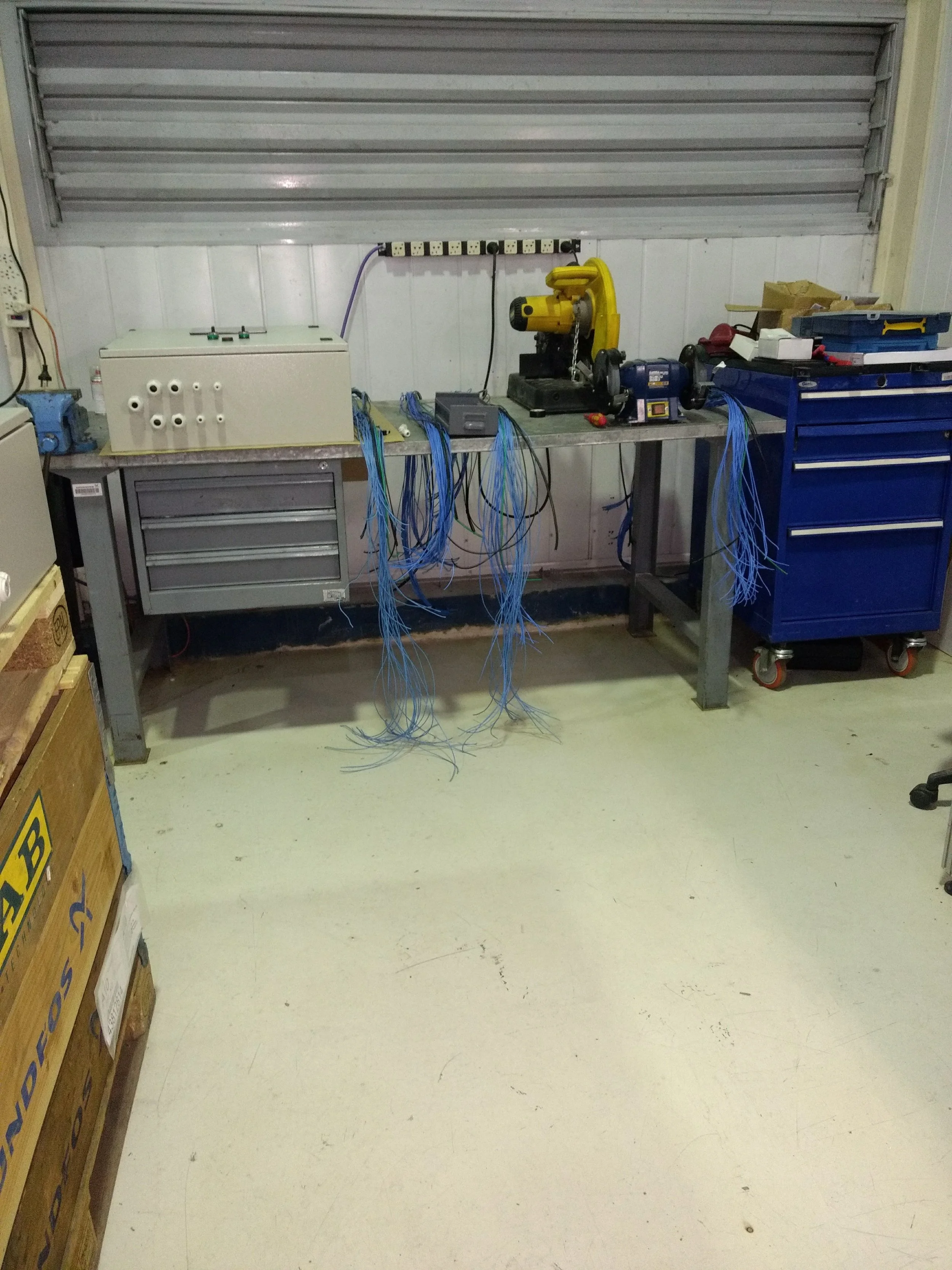 Un espacio de taller con una mesa de trabajo, máquinas eléctricas y herramientas, cables azules colgando y cajas de madera a la izquierda.