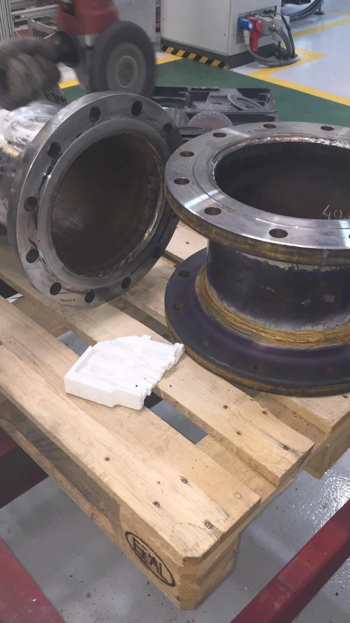 Componentes metálicos grandes, posiblemente partes de maquinaria, sobre una mesa de madera en una fábrica o taller, con maquinaria y herramientas en el fondo.
