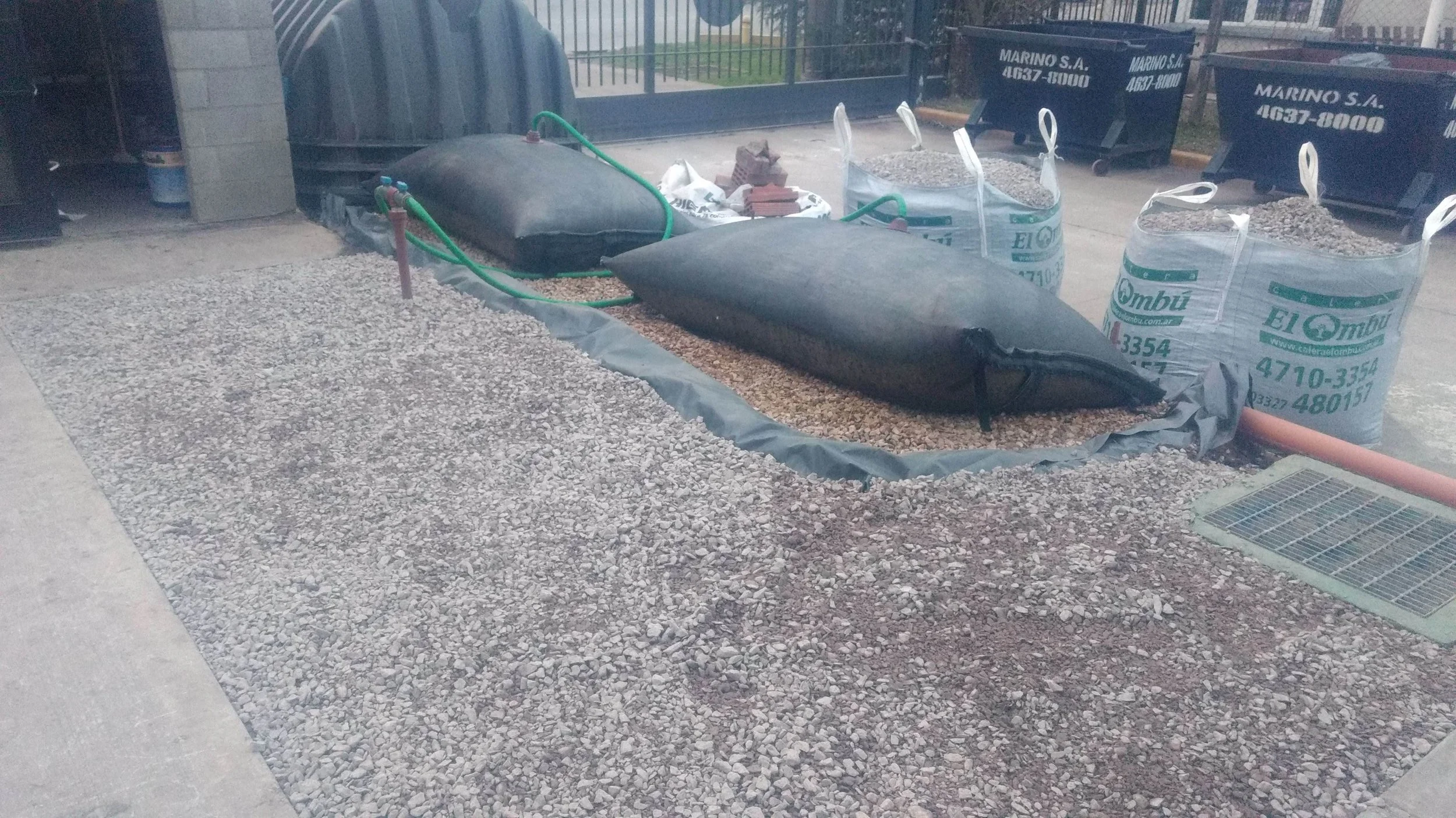 Área de construcción de jardín con fertilizante y herramientas, en un recinto con vallas de metal y botes de basura en el fondo.