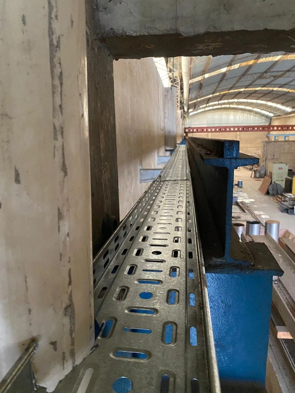 Vista de una estructura metálica en una fábrica o taller, con un techo alto y curvo, y algunas máquinas y materiales en el fondo.
