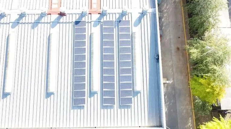 Paneles solares instalados en el techo de un edificio de chapa metálica, con árboles y una acera al lado.